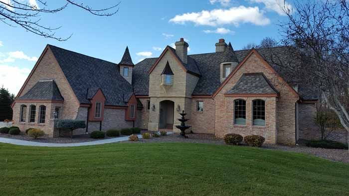 Large brick house with steep roofs, arched entry, and a small fountain in the front yard under a blue sky