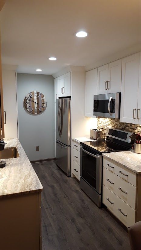Bright narrow kitchen with white cabinets, stainless appliances, dark floor, and recessed ceiling lights.