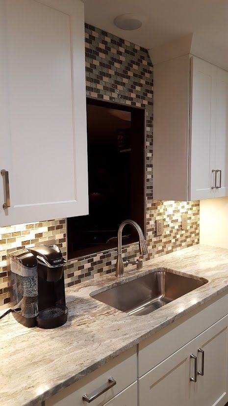 Modern kitchen counter with sink, chrome faucet, coffee maker, white cabinets, and tiled backsplash over a pass-through window