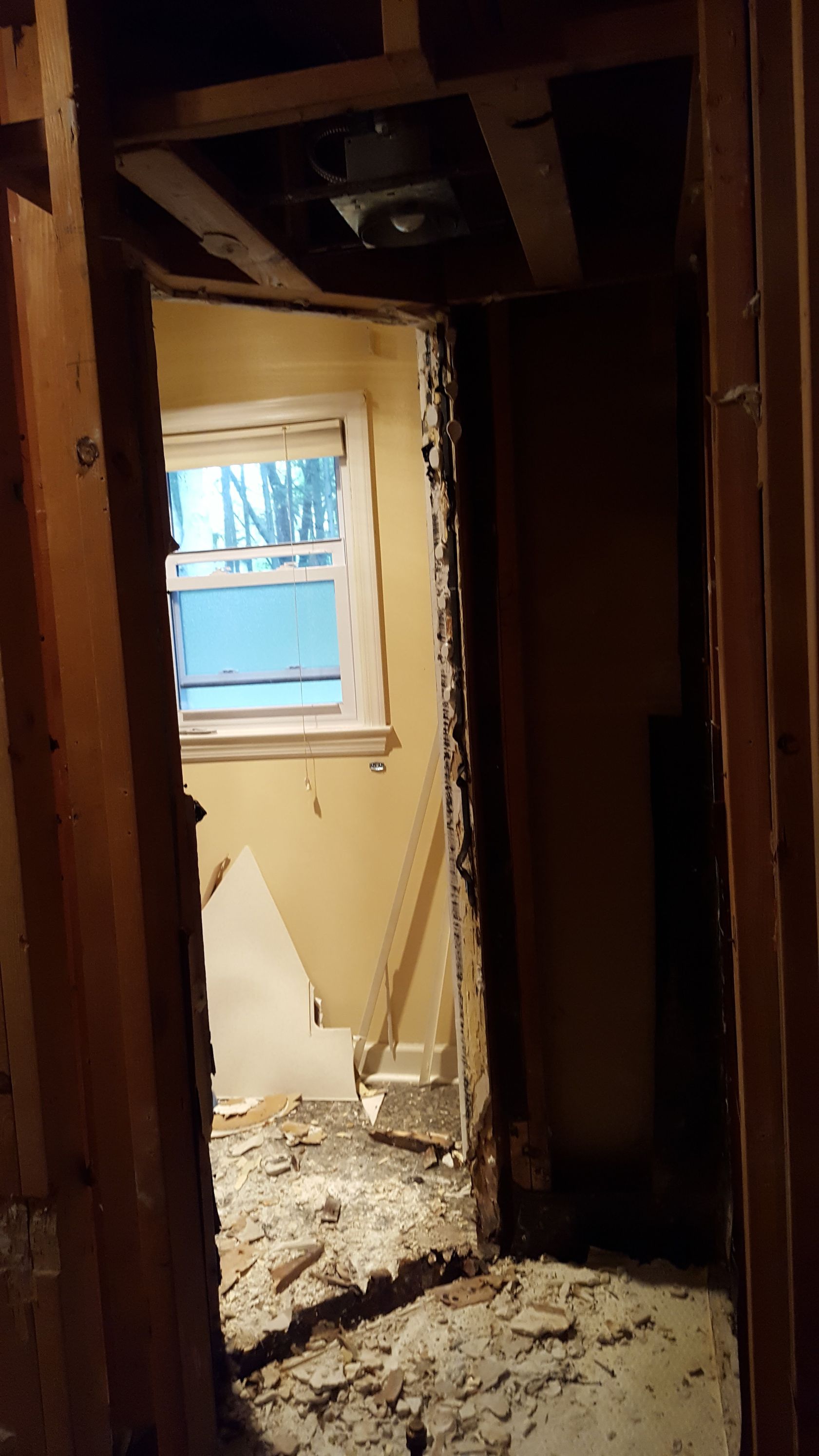 Damaged hallway with exposed studs, debris on the floor, and a small window at the end