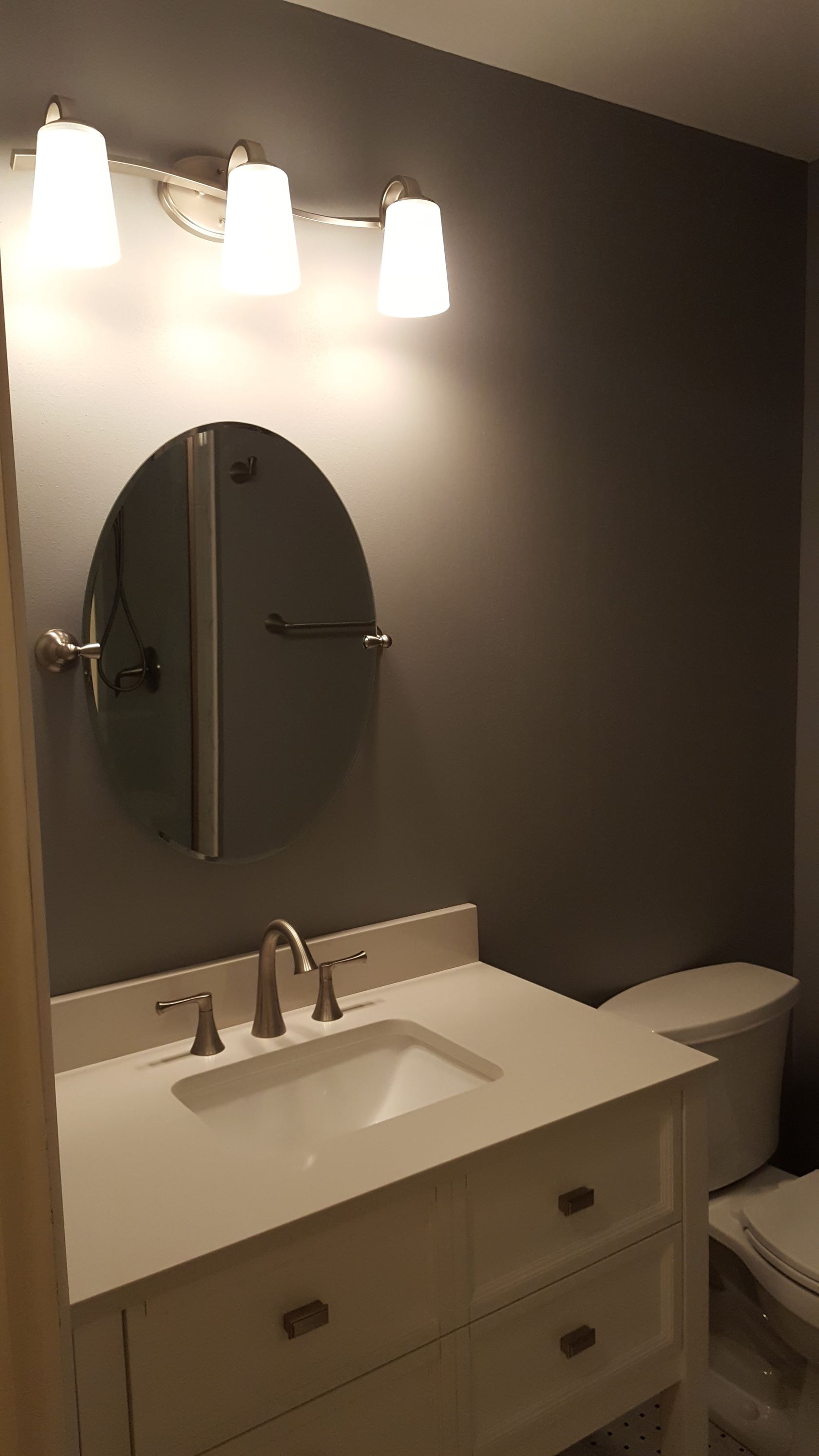 Bathroom vanity with white sink, round mirror, three wall lights, and a toilet beside it