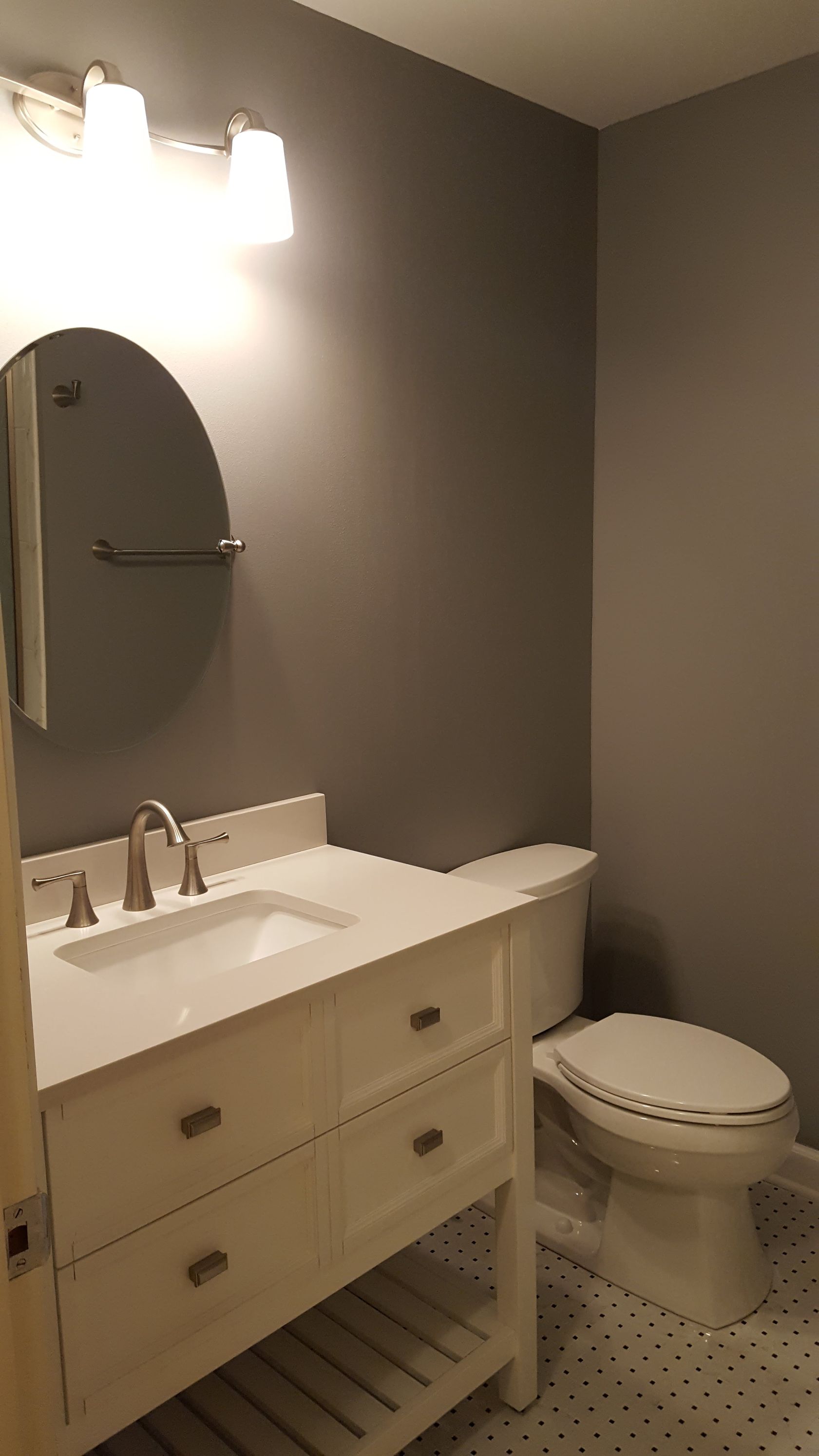 Modern bathroom with white vanity, oval mirror, sink, toilet, and gray walls.