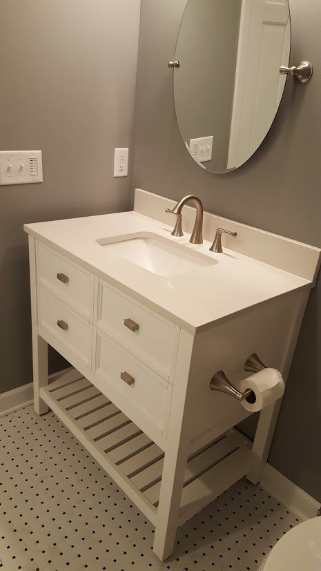 White bathroom vanity with sink, mirror, and wall-mounted faucet in a gray tiled restroom.