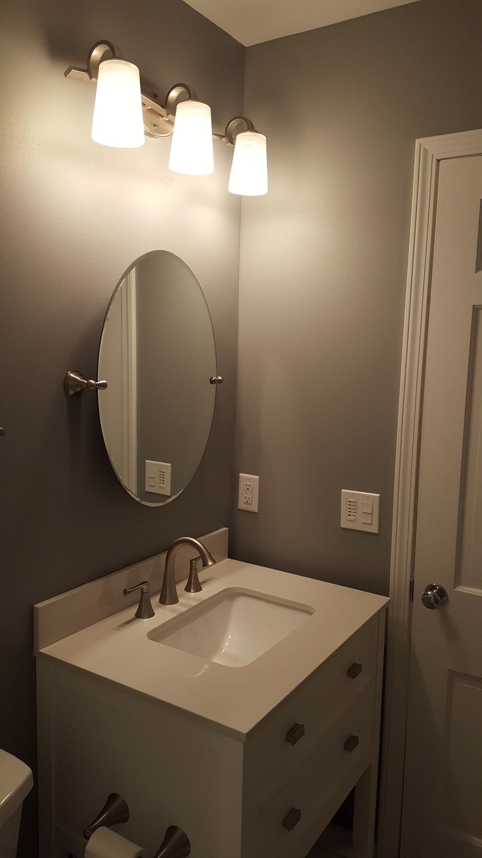 Small bathroom vanity with oval mirror, white sink, gray walls, and three pendant lights.