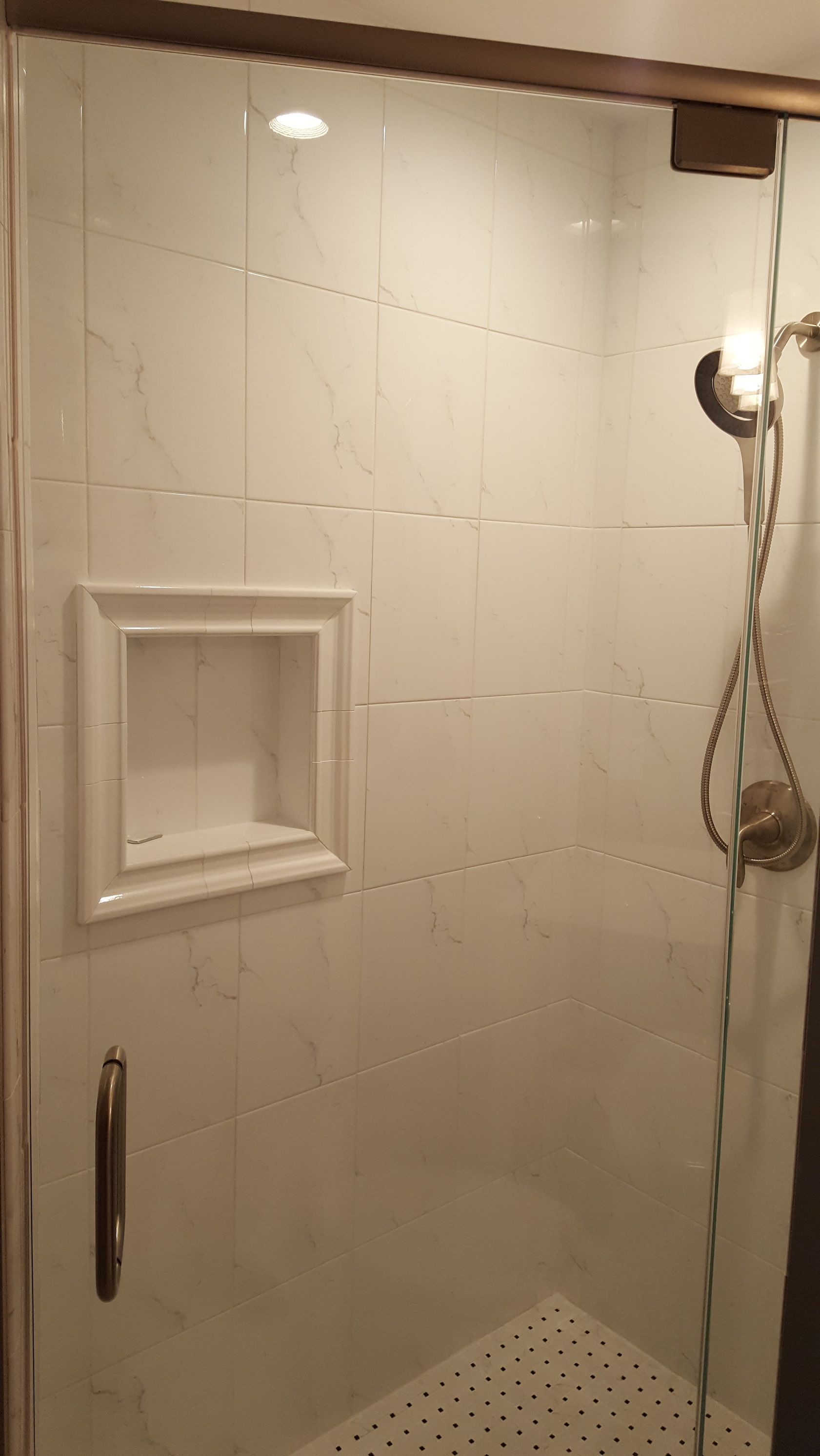 White-tiled shower stall with built-in shelf, handheld showerhead, and glass door.
