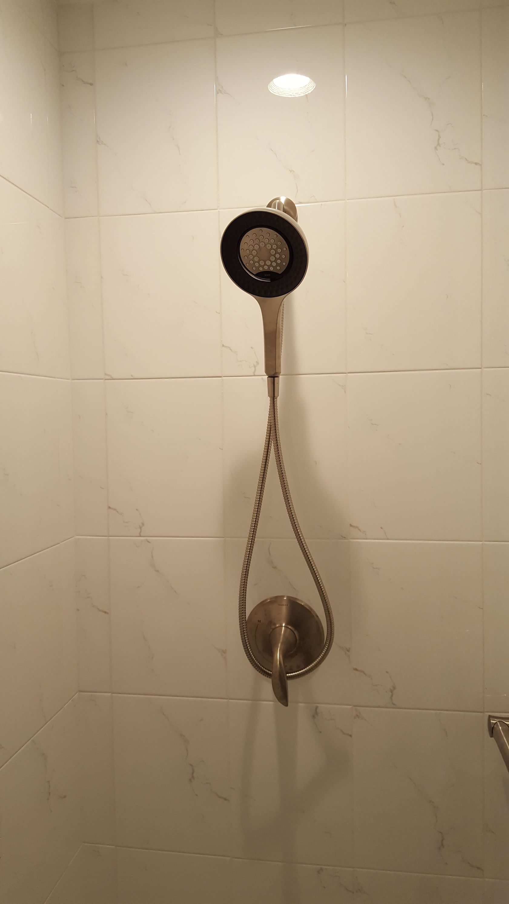 Wall-mounted showerhead with a hanging handheld shower in a white-tiled bathroom.