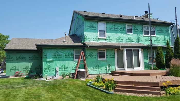 Green two-story house with deck, ladder, and a lawn under a clear blue sky