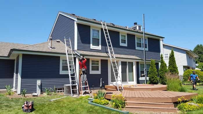 Workers on ladders repainting a blue two-story house with a wooden deck in front