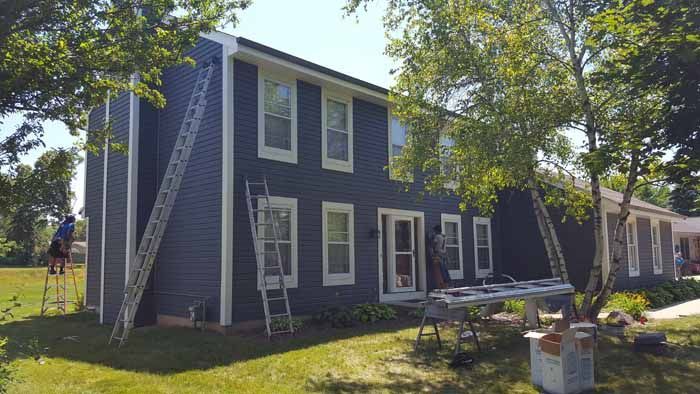 Two-story blue house with white trim, ladders leaning against it, and tools in the yard.