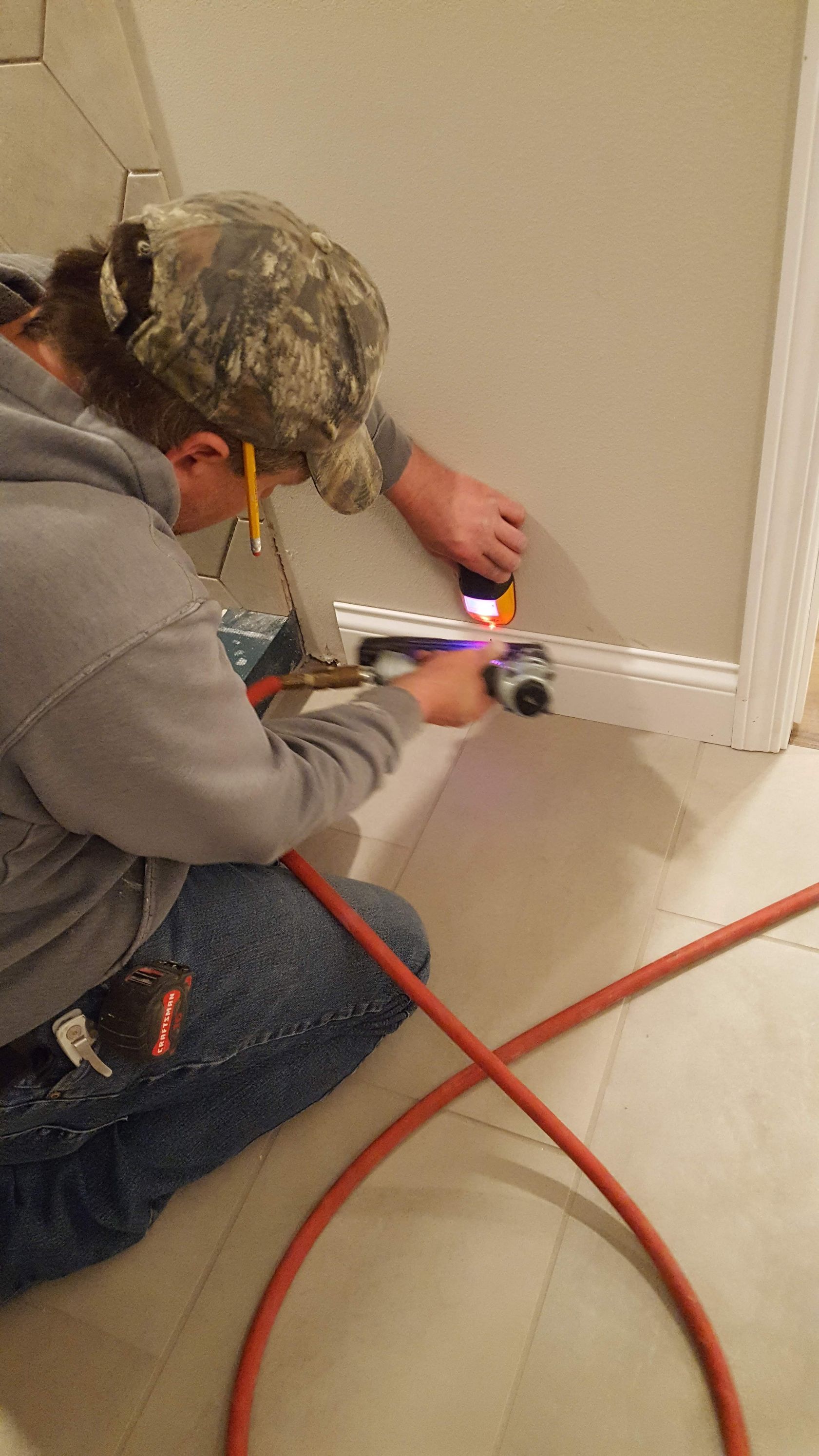 Person kneeling with a power tool, cutting drywall near a white baseboard in a room.