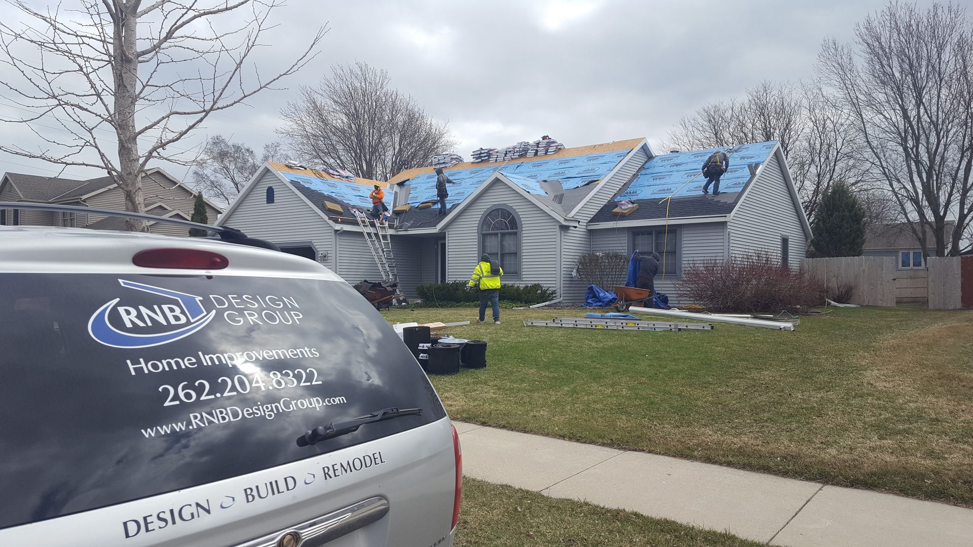 Workers repair a storm-damaged house with blue tarps on the roof, viewed from a van in front.