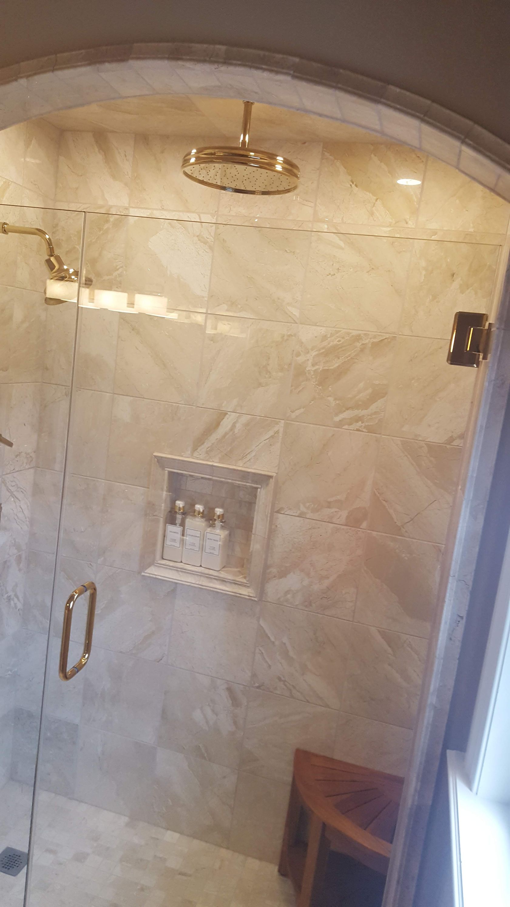 Glass shower with beige stone tile, rainfall showerhead, and a small wooden stool inside.