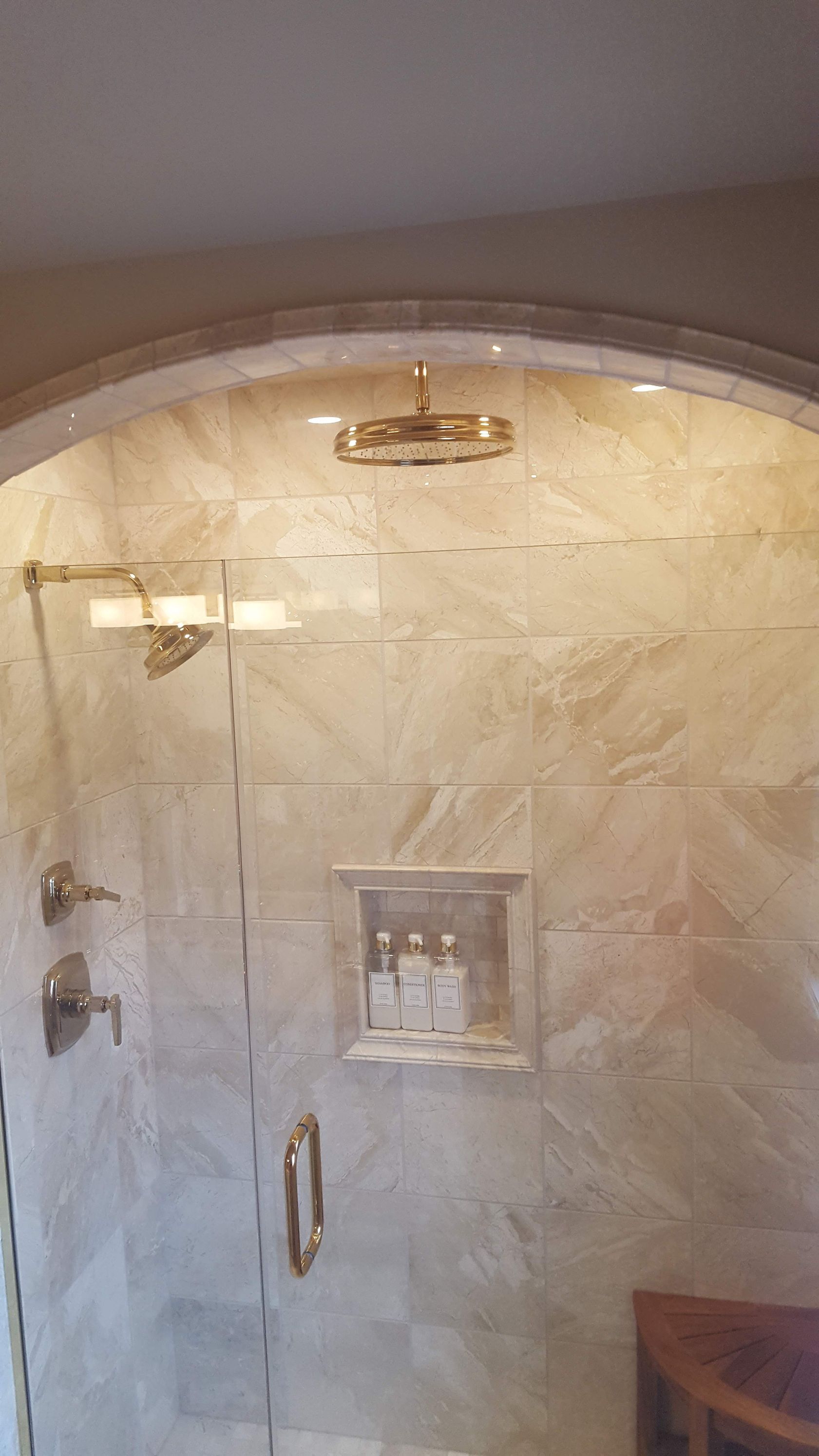 Bathroom shower with beige tile, glass door, and brass fixtures under an arched entryway