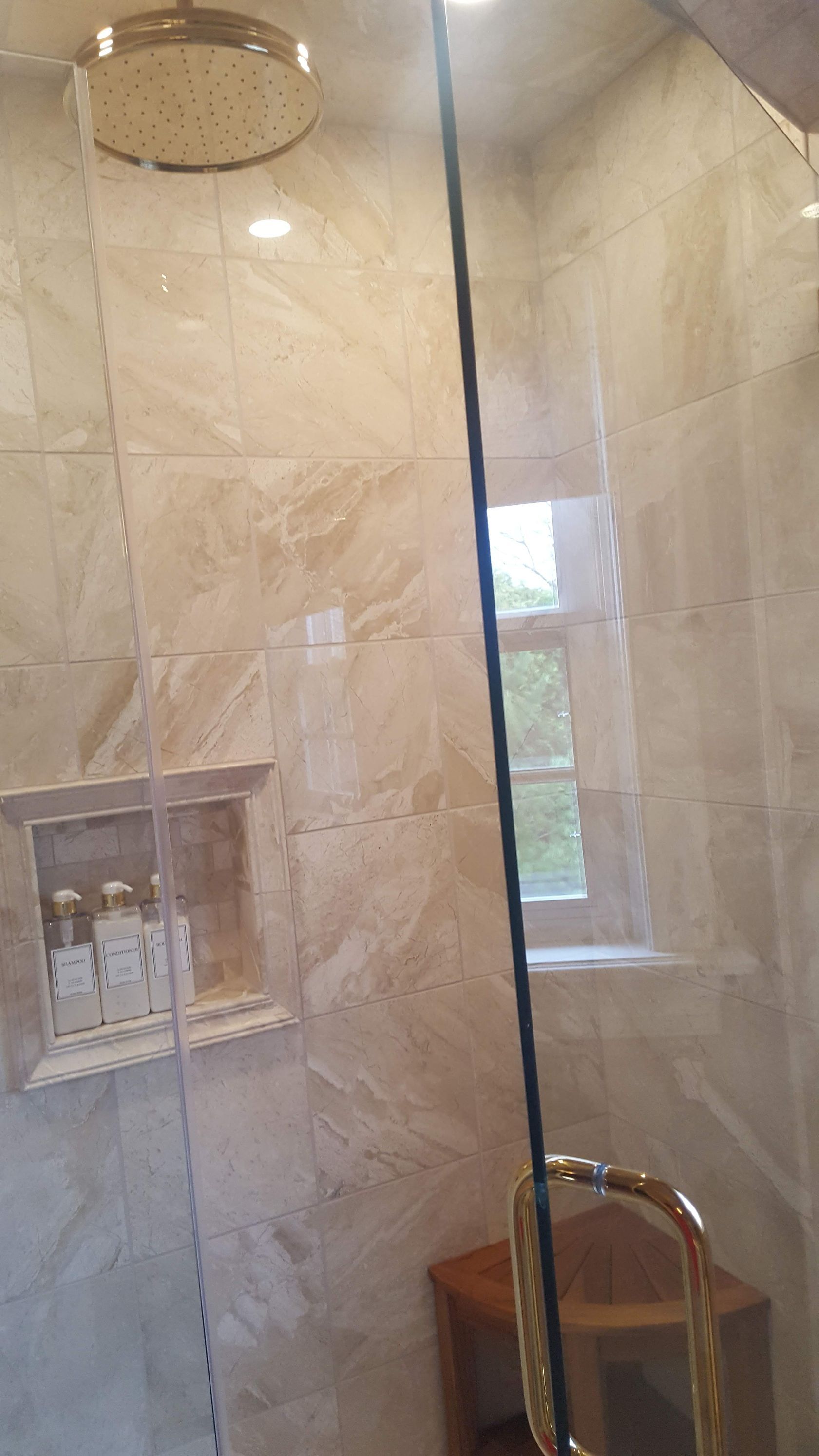 Glass shower with beige tile walls, round overhead showerhead, and a small built-in shelf.
