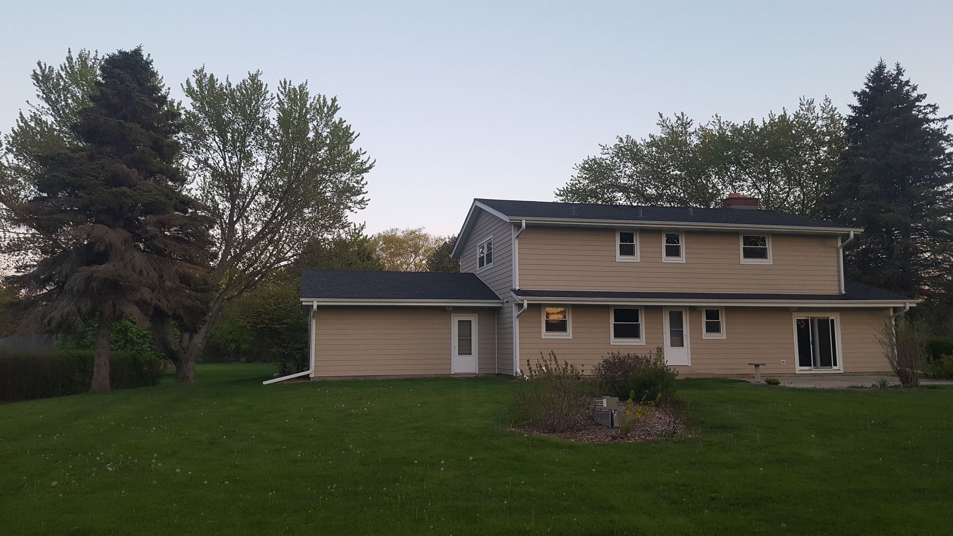 Two-story beige house with a large green lawn and trees in the front yard at dusk