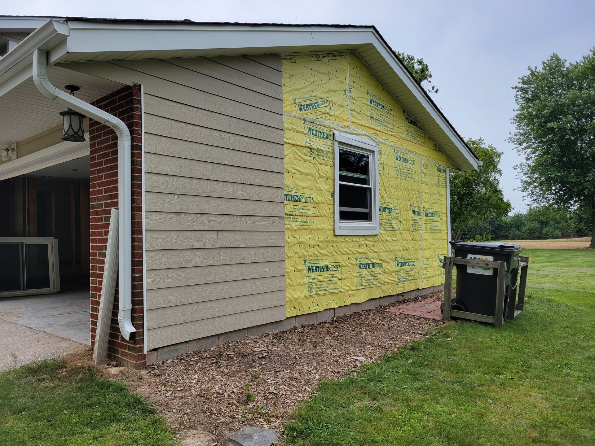 Yellow house exterior with tan siding and exposed construction wrap beside a grassy yard