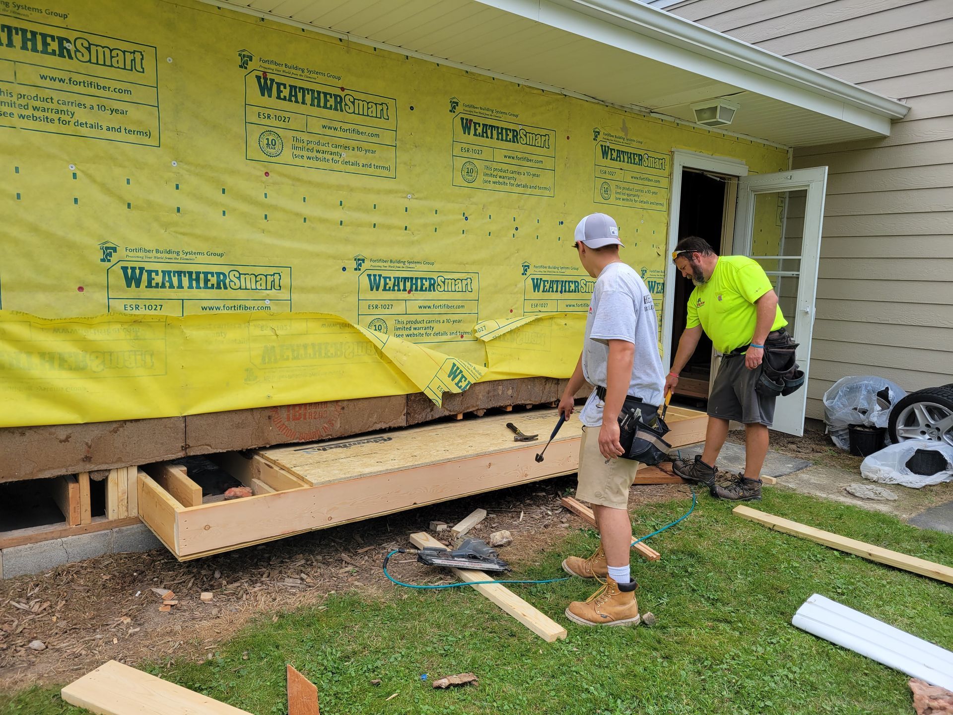 Two workers inspect house siding under construction with yellow house wrap exposed around the foundation.