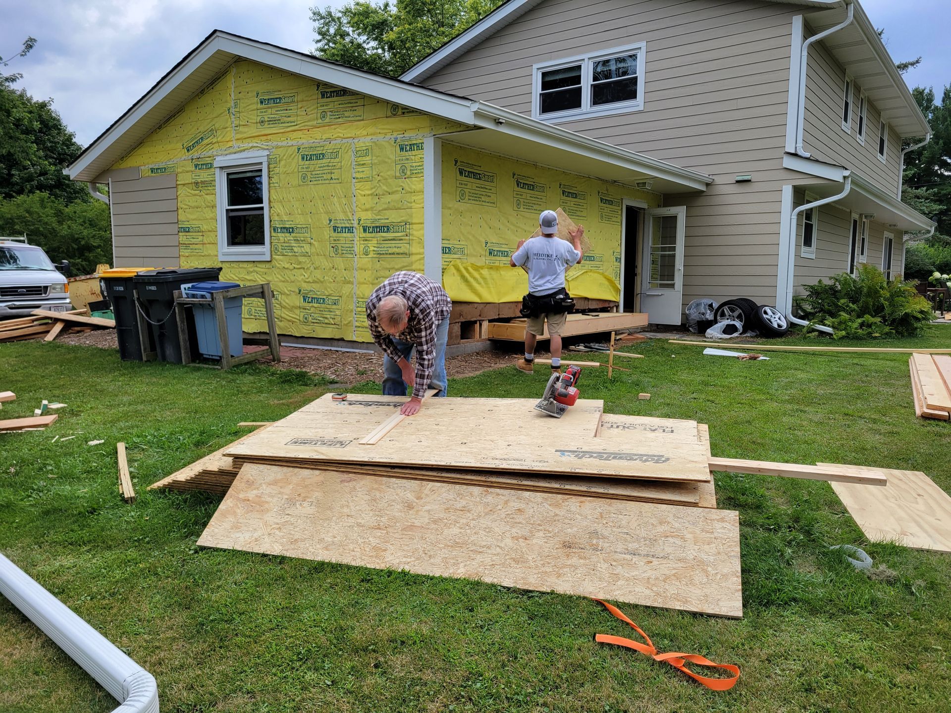 Workers installing plywood on a house exterior with green grass and yellow siding