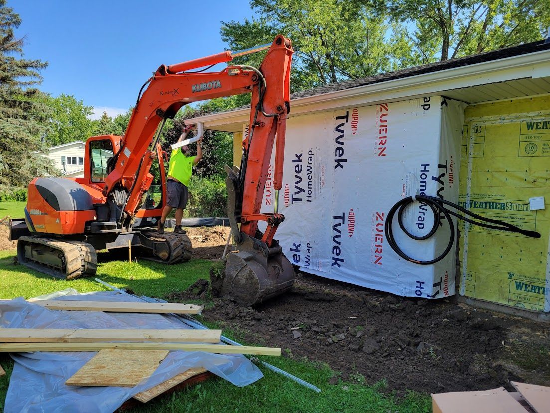 Orange excavator digs beside house under construction with wrapped wall panels and a worker nearby