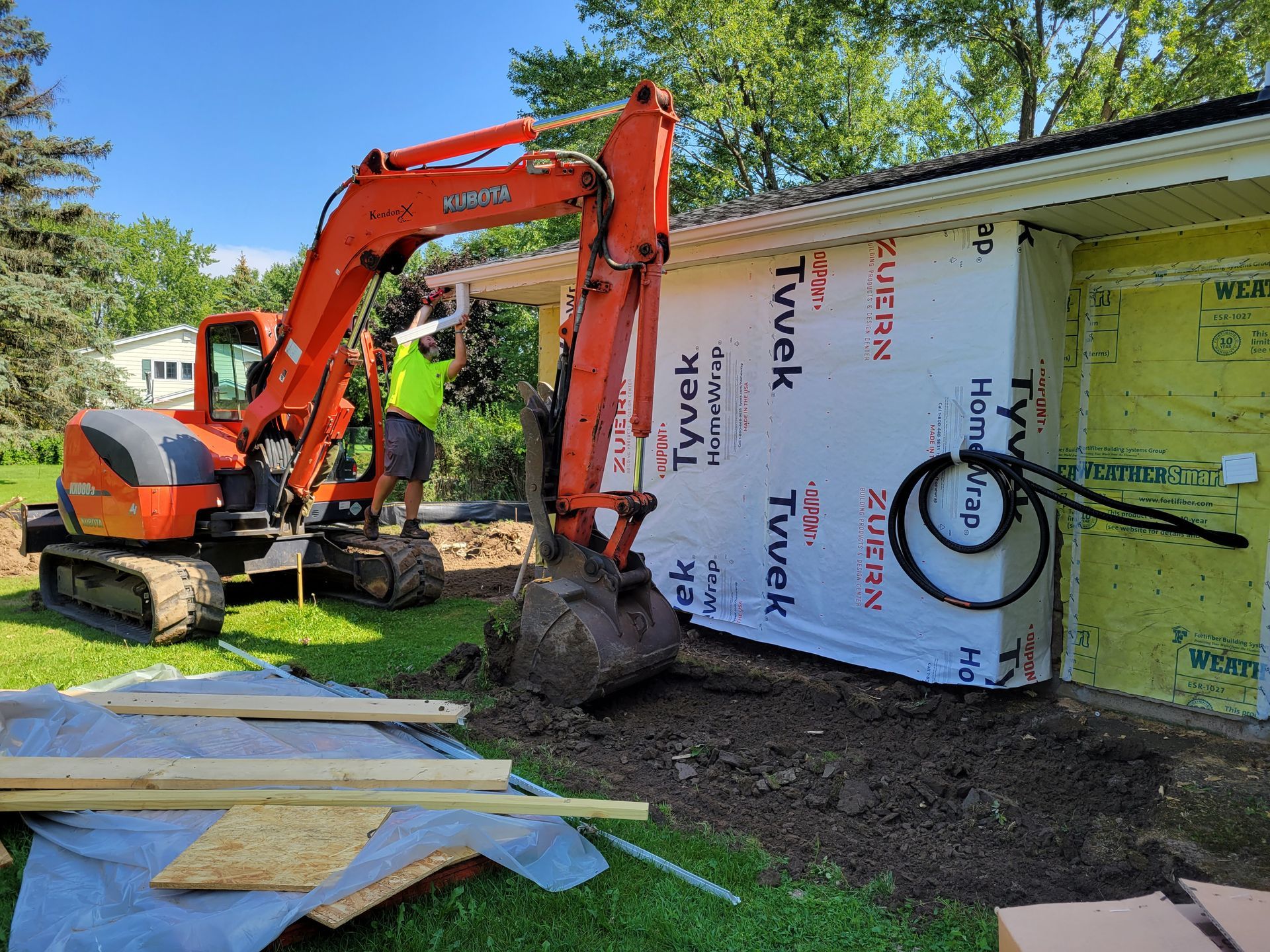 Orange excavator beside a house under construction, with workers and building wrap exposed.