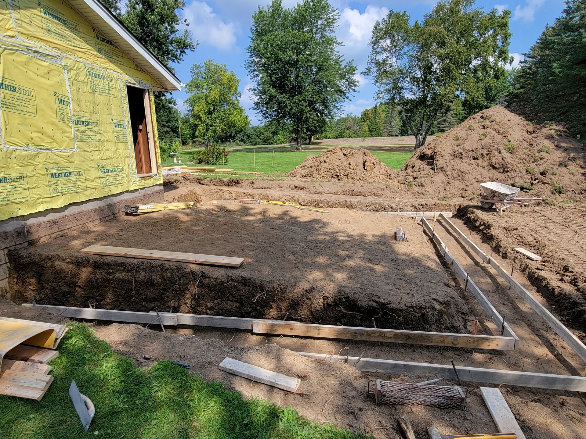 Partially excavated yard beside a yellow house, with dirt piles, wooden forms, and a work area in progress