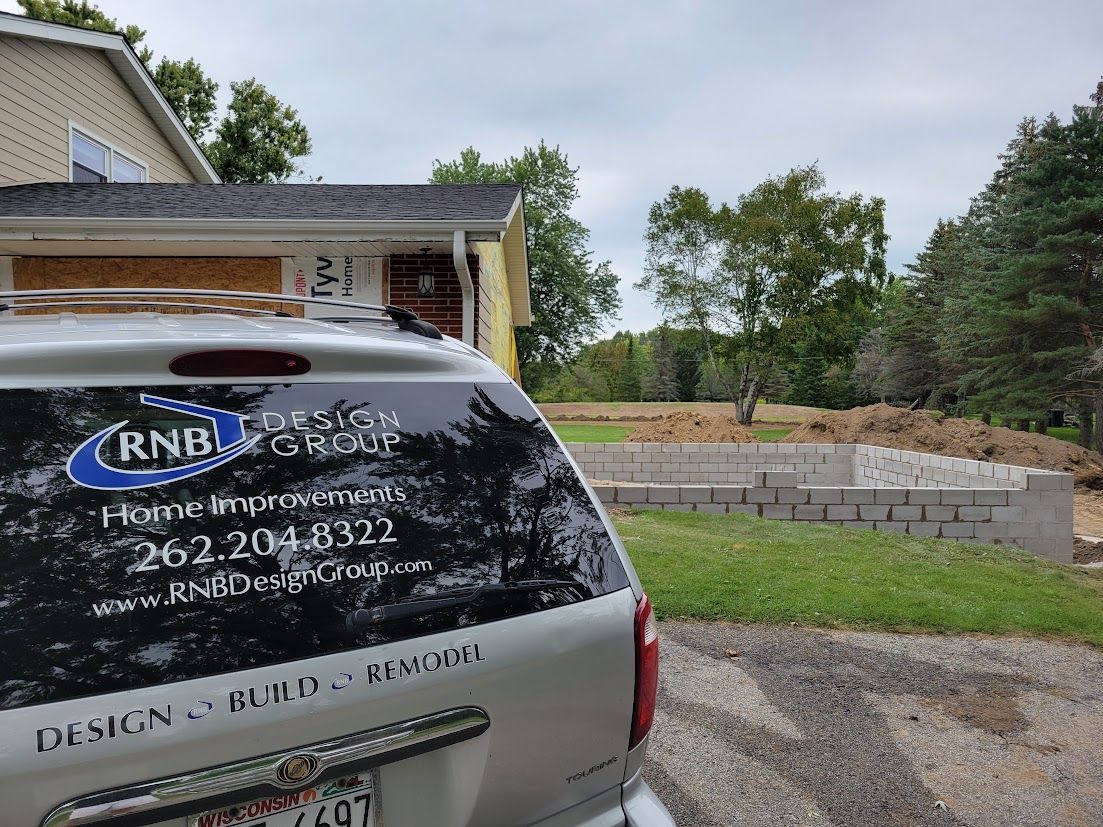 Silver SUV with RNB decal parked beside a house and stone retaining wall on a gravel driveway
