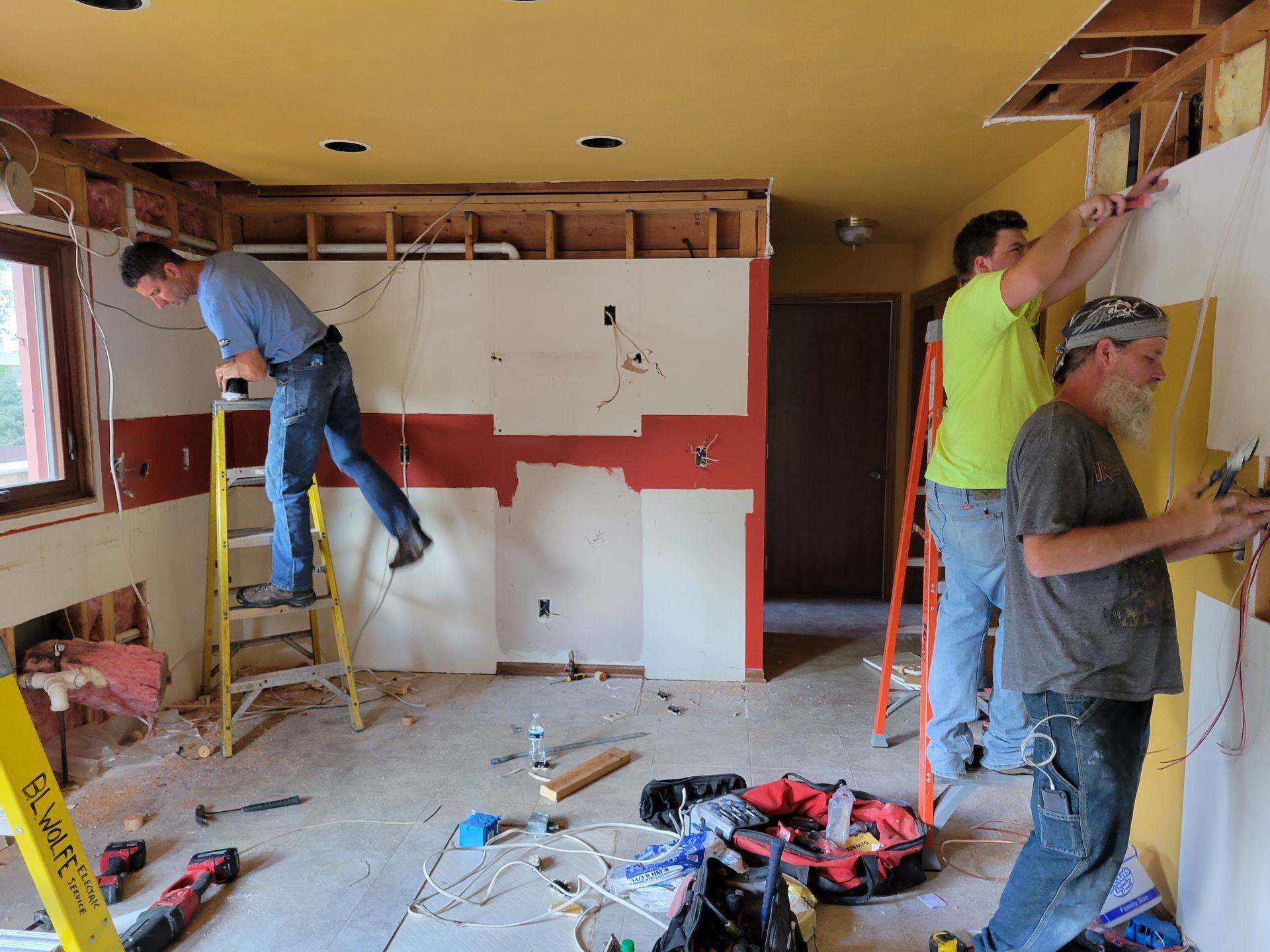 Workers renovating a room, one on a ladder and two at a wall with exposed framing and drywall.