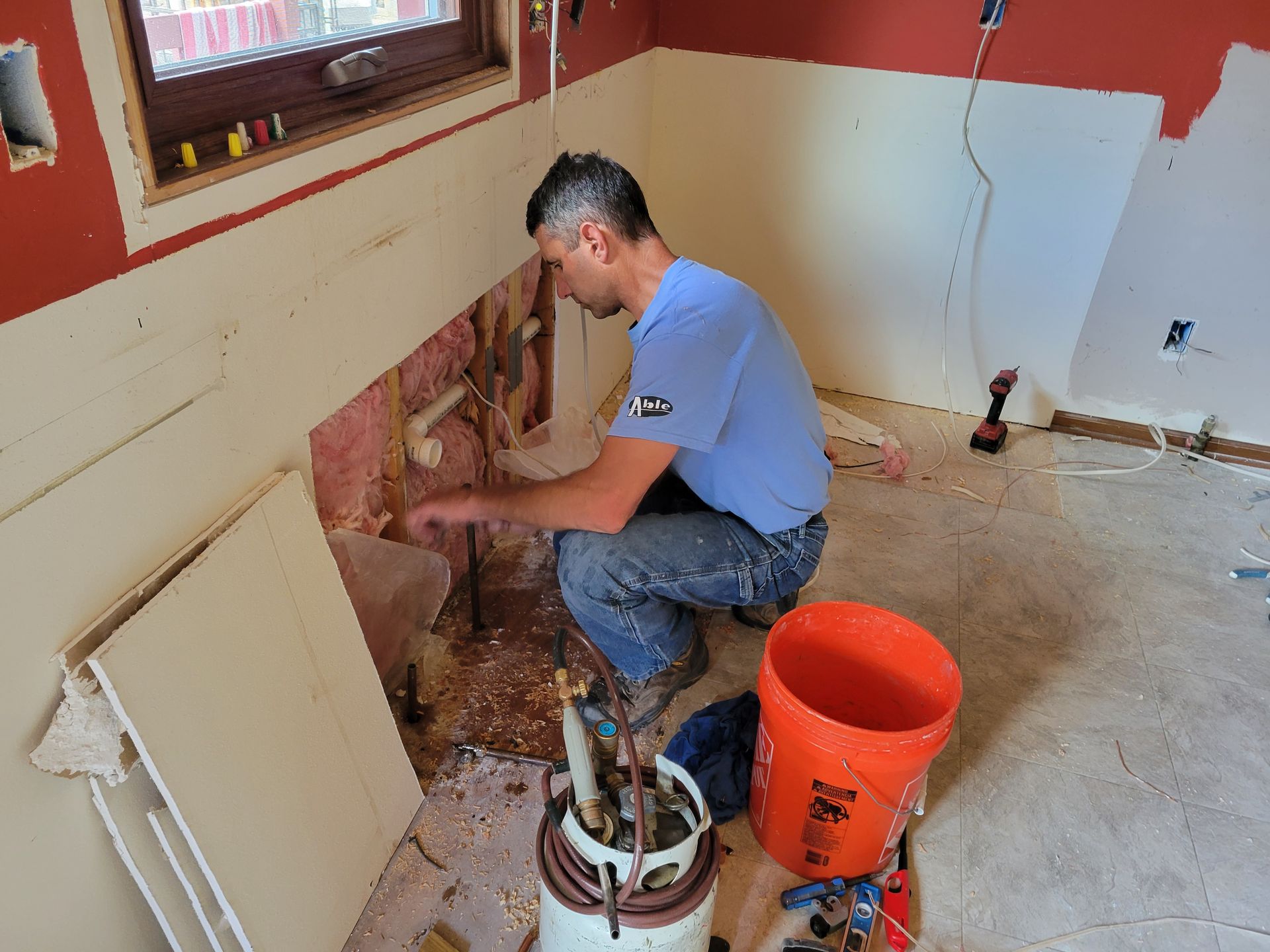 Person kneeling by a wall cutout, doing home repair amid tools, buckets, and exposed plumbing