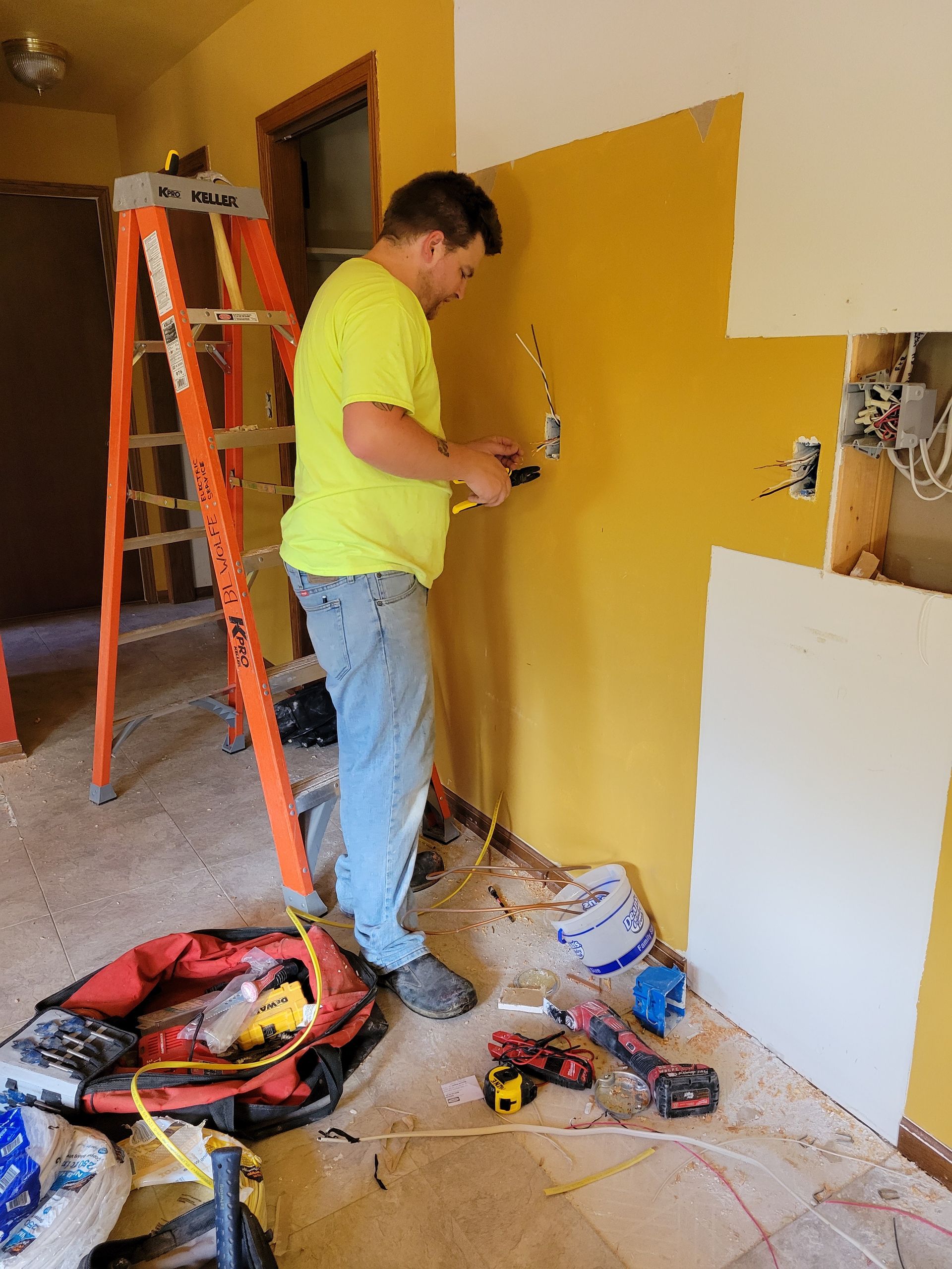 Person painting a yellow wall in a room with tools, ladder, and supplies on the floor
