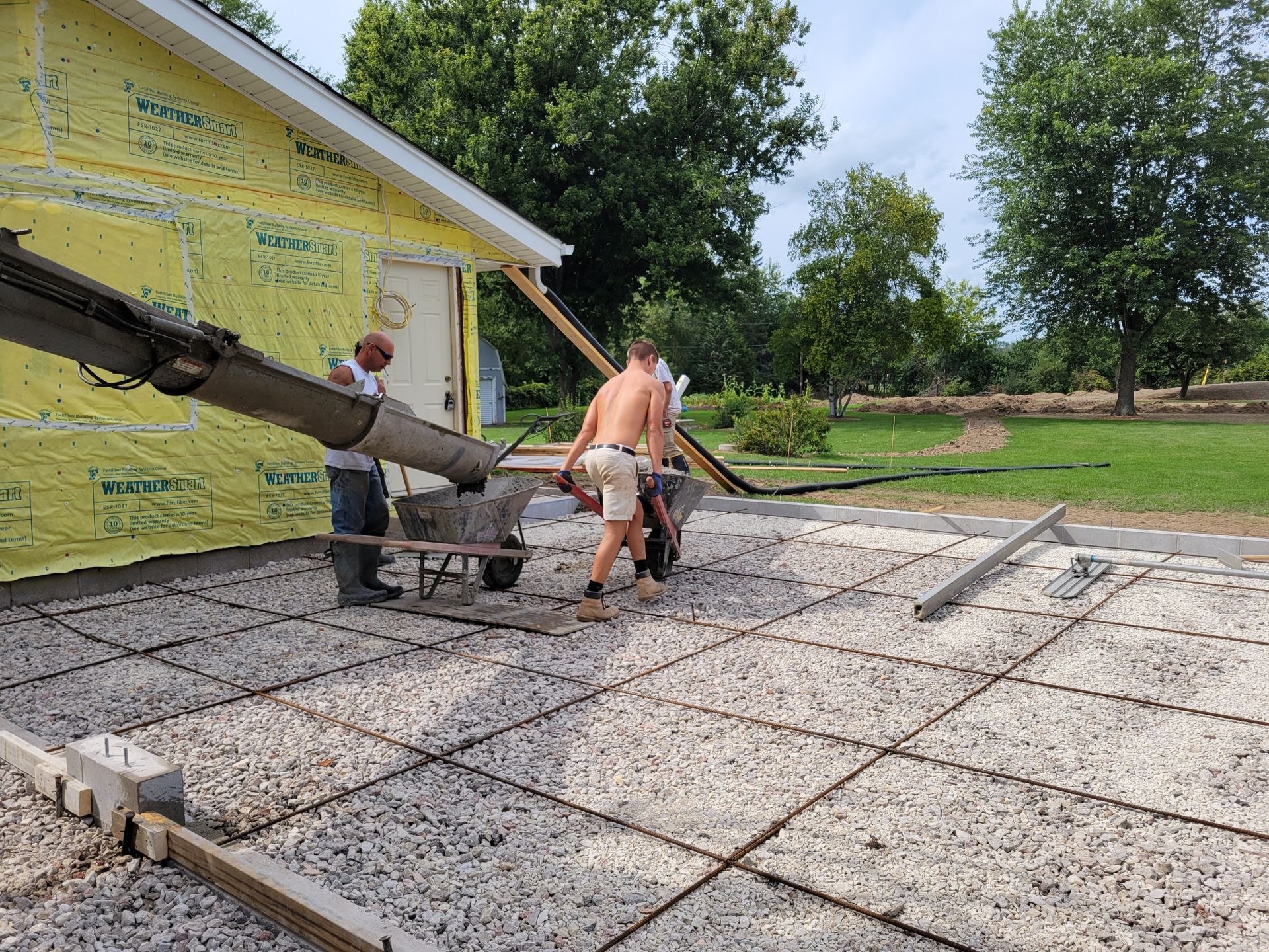 Construction workers pouring a concrete slab beside a yellow house, with trees and yard in the background