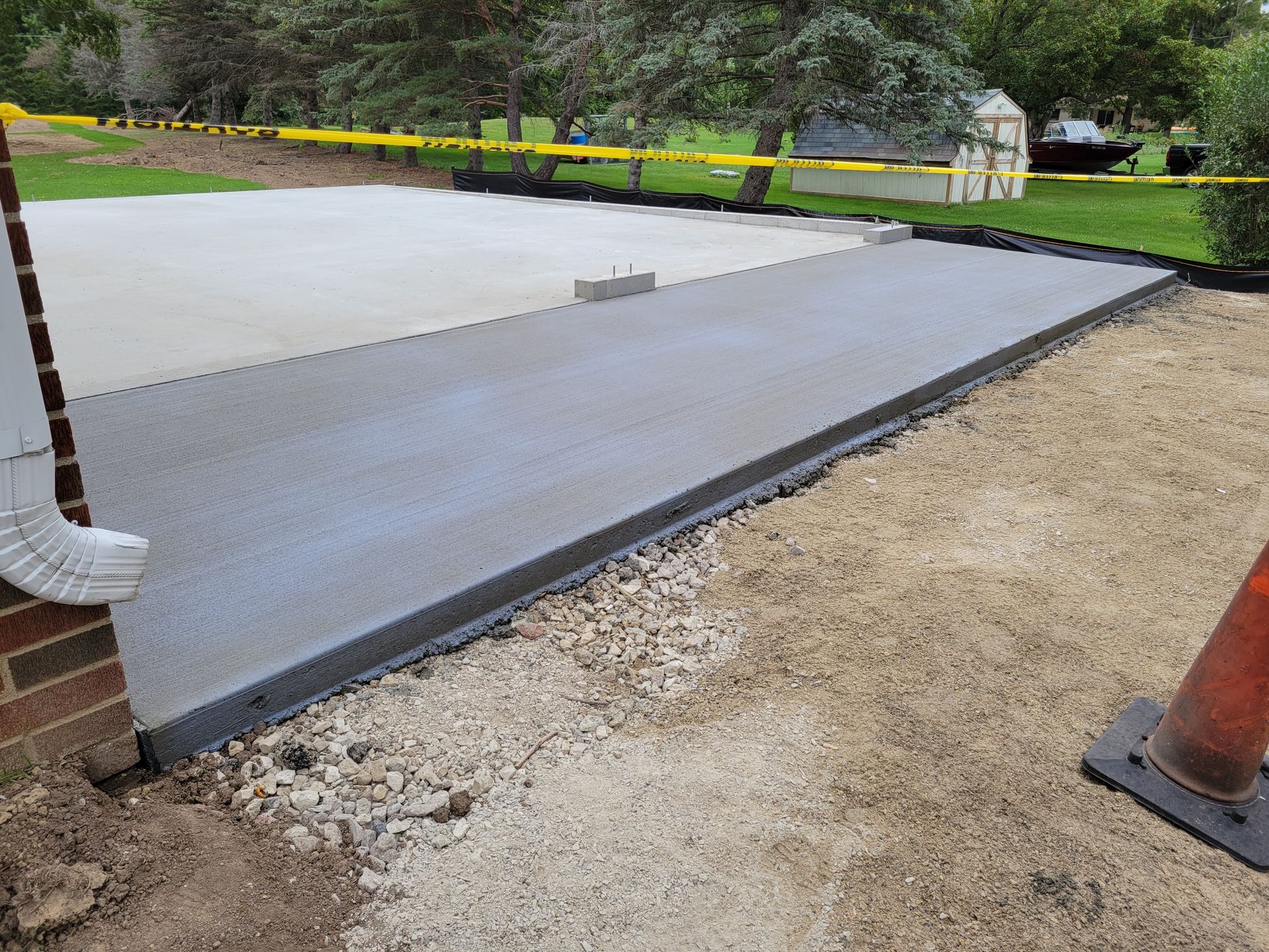Freshly poured concrete driveway apron beside a gravel path, with caution tape and a wooded yard.