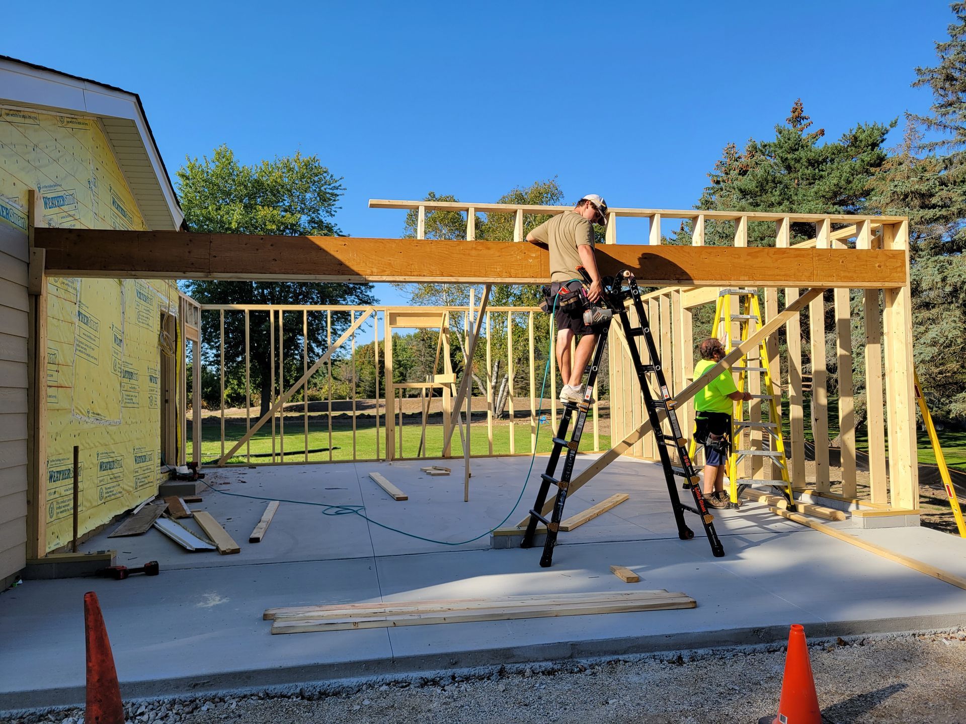 Construction workers framing a house addition with ladders, lumber, and tools on a concrete slab.