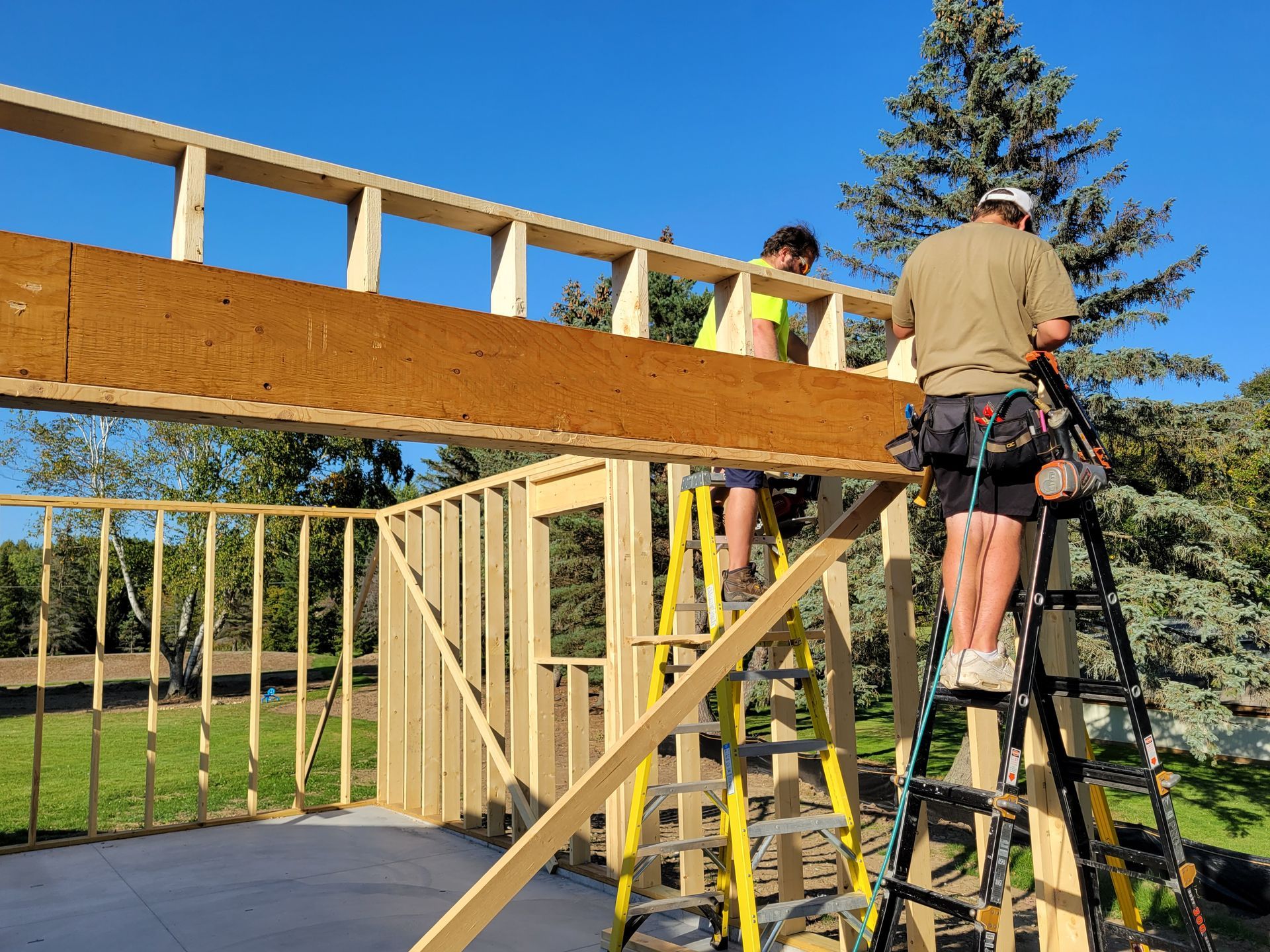 Workers building a wooden deck frame outdoors beside a ladder and trees