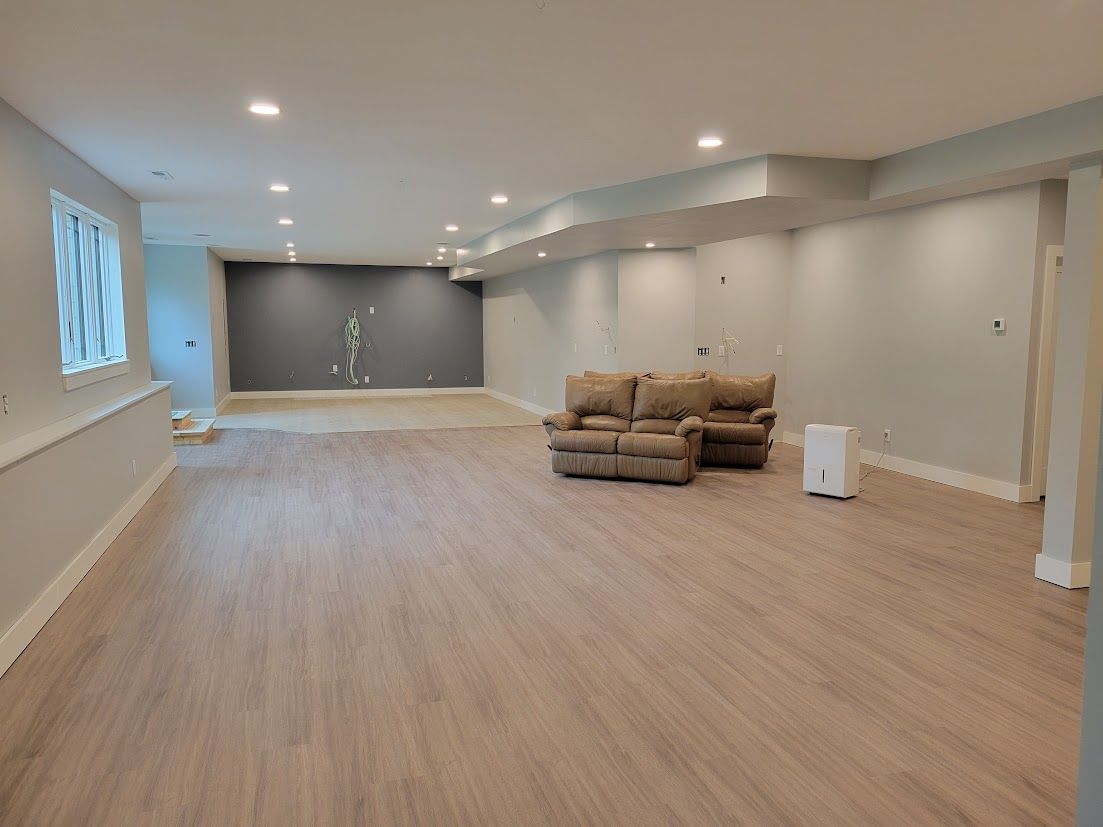 Empty finished basement with light wood floors, white walls, recessed lights, and a small gray seating area.