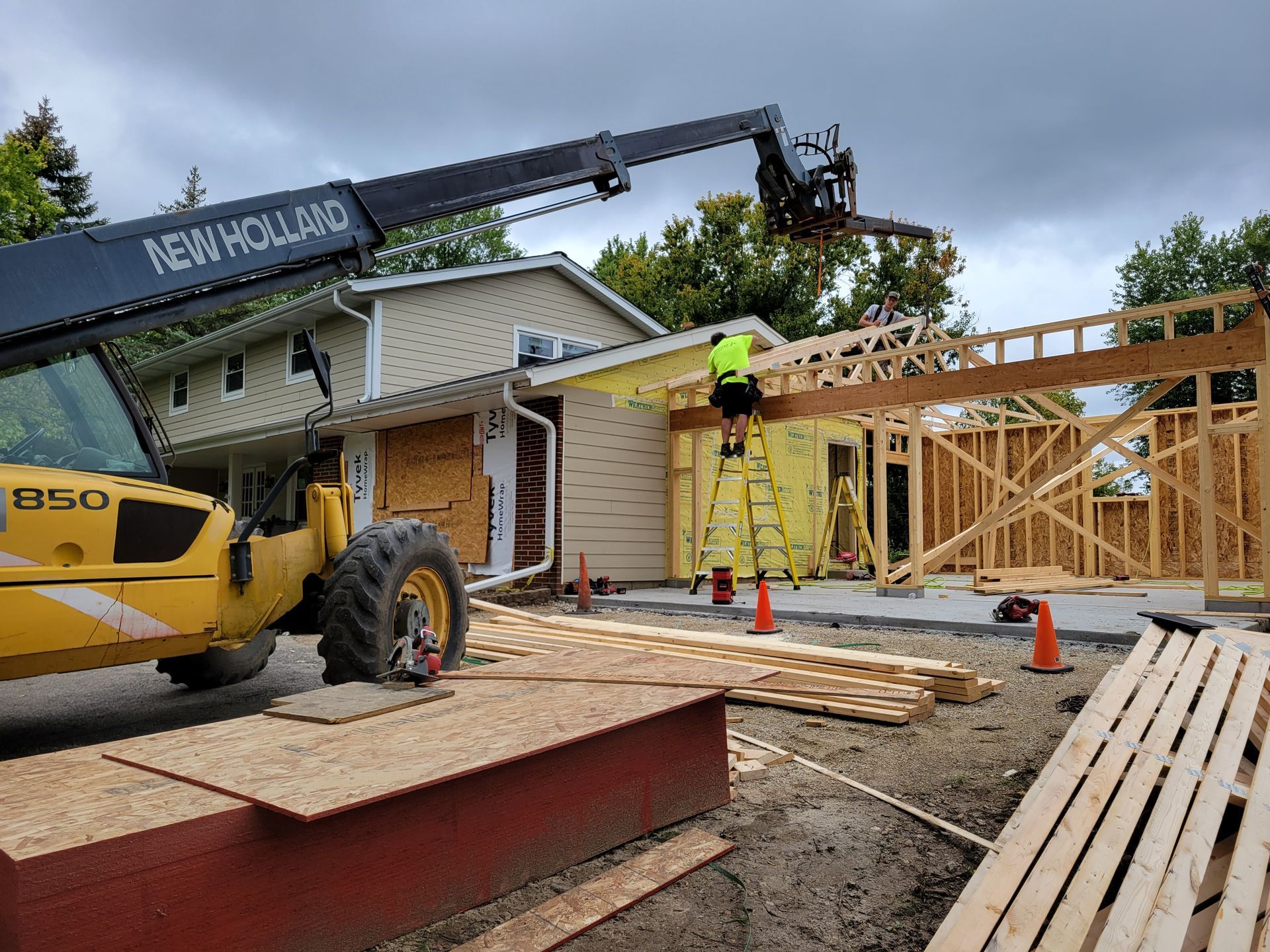 Construction crew framing a house addition beside a yellow excavator and crane under a partly cloudy sky