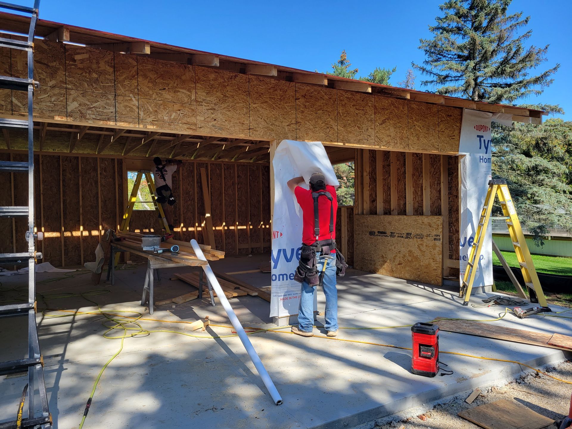 Workers framing a house addition under construction, with ladders, tools, and a concrete slab.