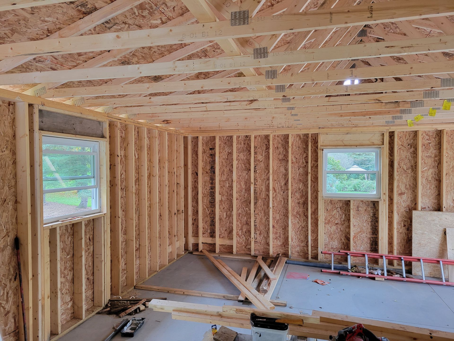 Unfinished room with exposed wooden framing, two windows, and construction materials on the floor.