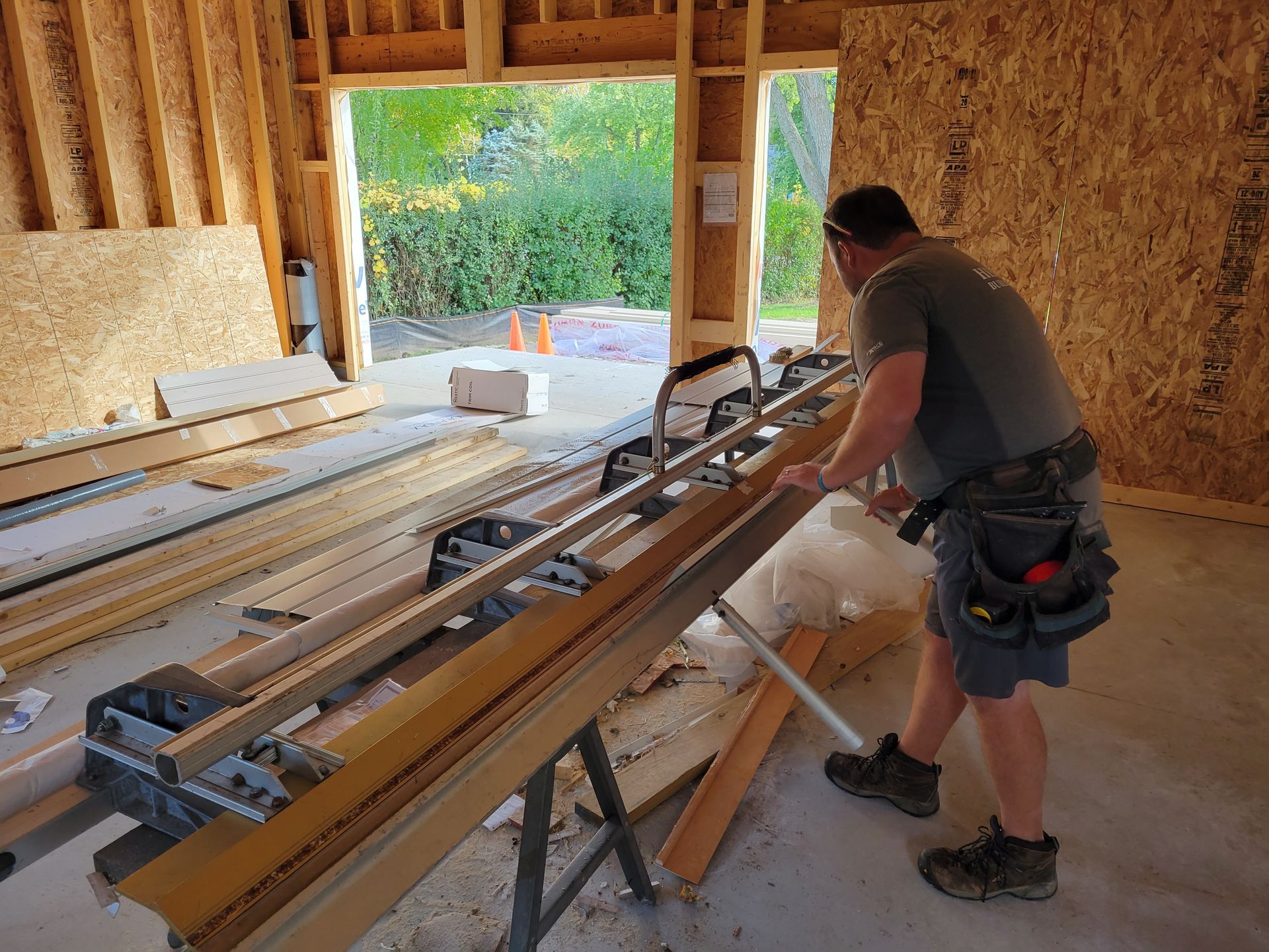 Worker operating a table saw in an unfinished plywood-framed workshop, with lumber stacked nearby.