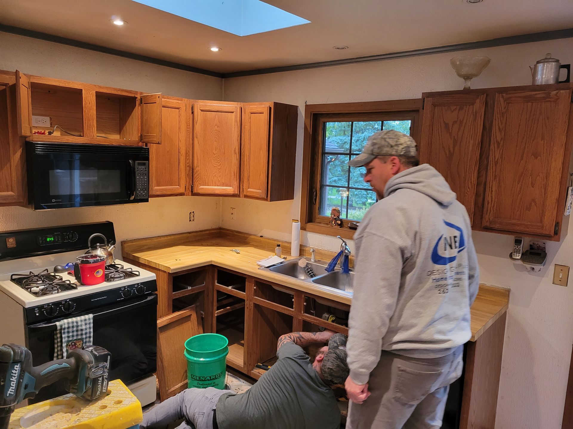 Person in gray hoodie standing in a kitchen beside a sink and cabinets, looking at a seated person.