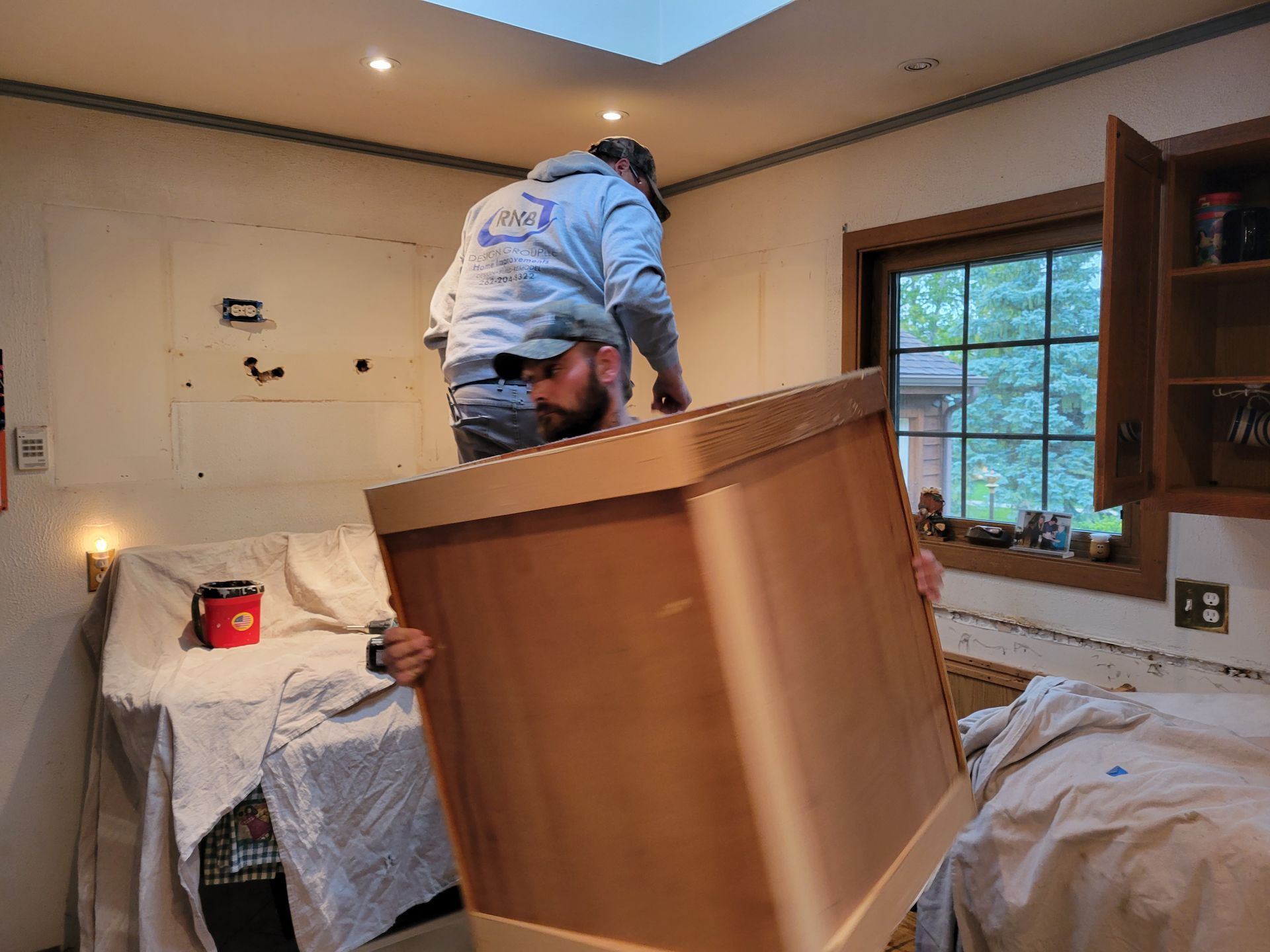 Person moving a large wooden cabinet in a cluttered bedroom with a bed and window.