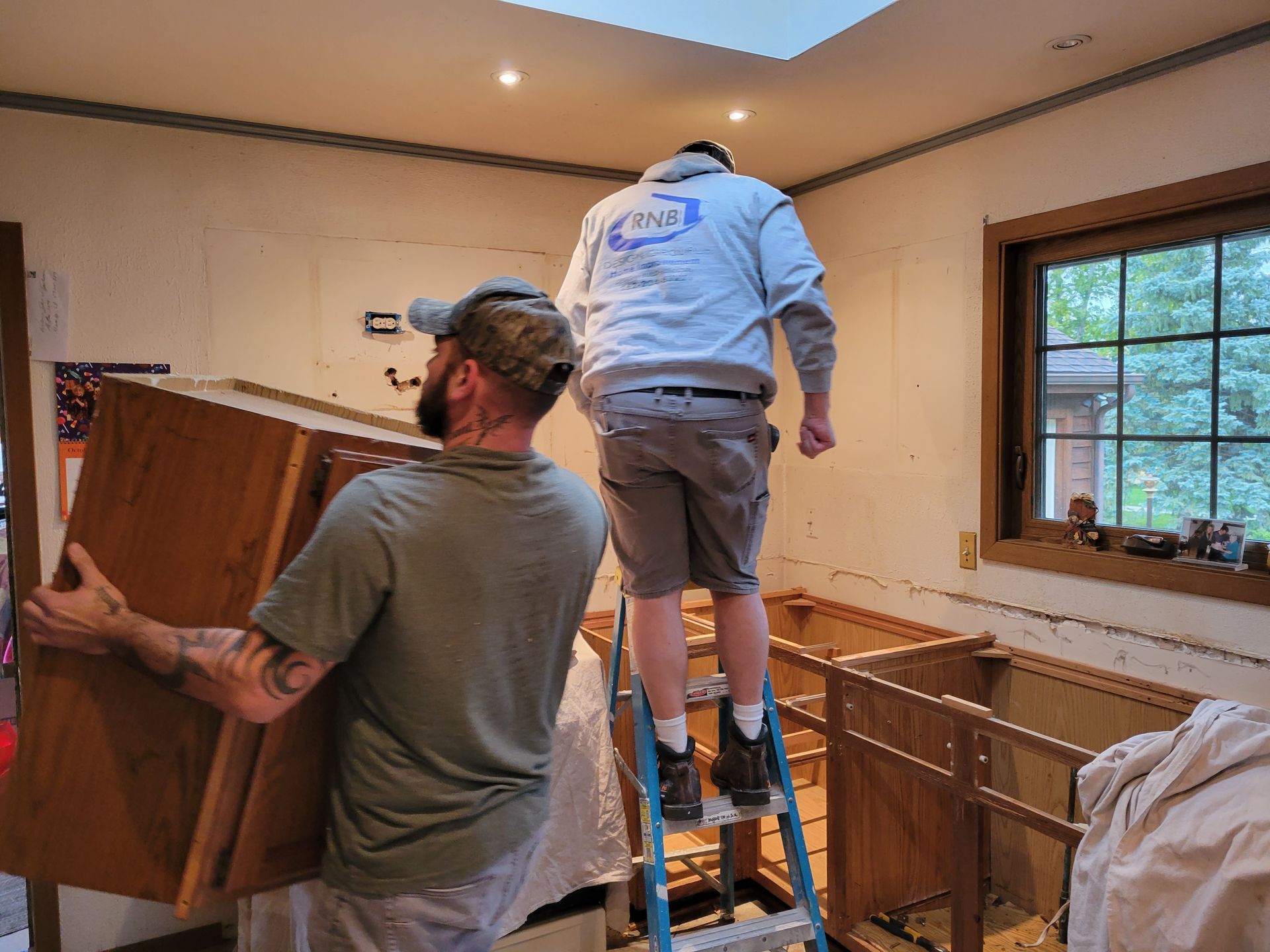 Two workers moving a large cabinet in a room under renovation, one standing on a step stool.