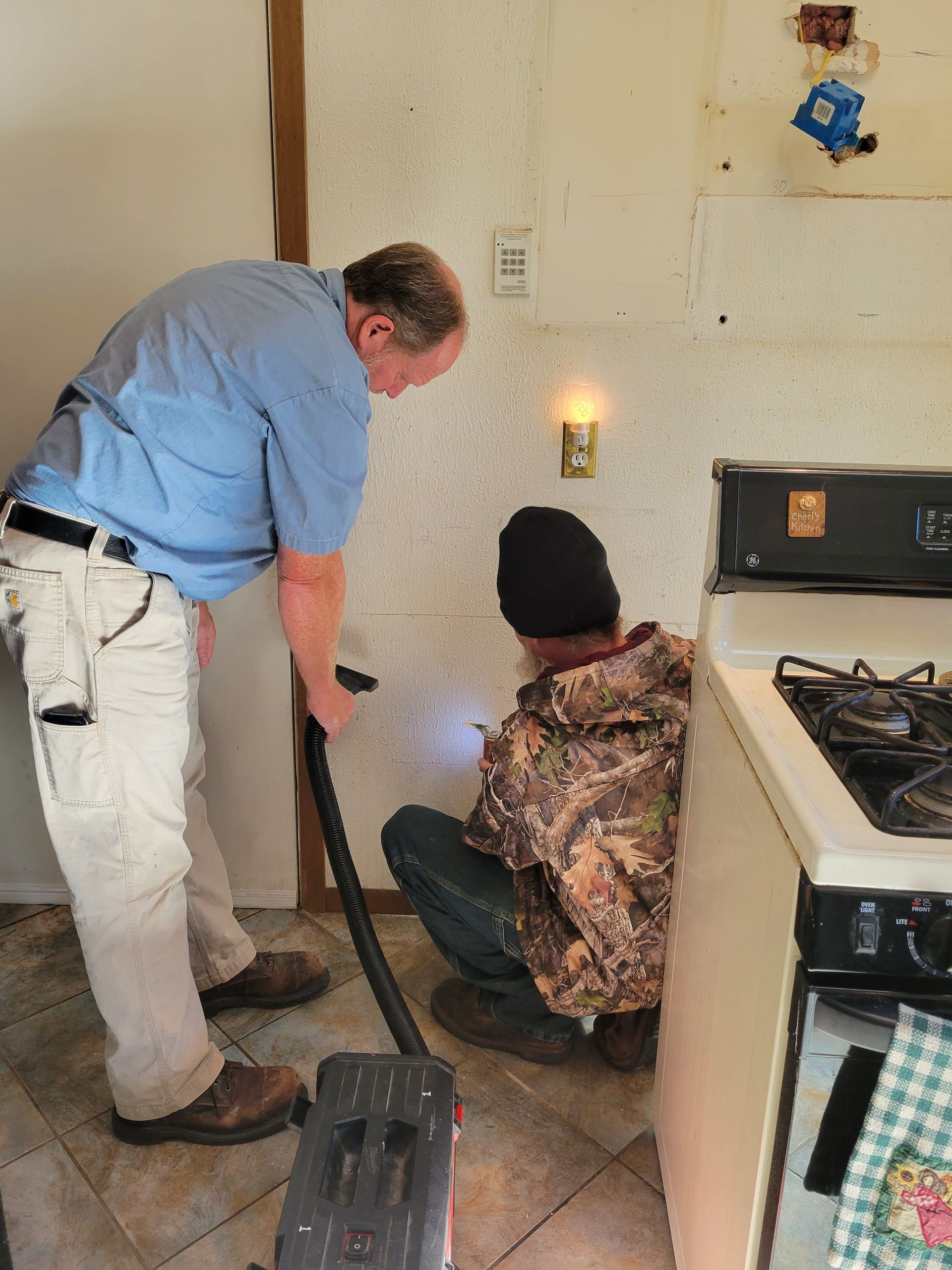 Two workers inspect a wall leak in a kitchen, one holding a vacuum hose near a lit hole.