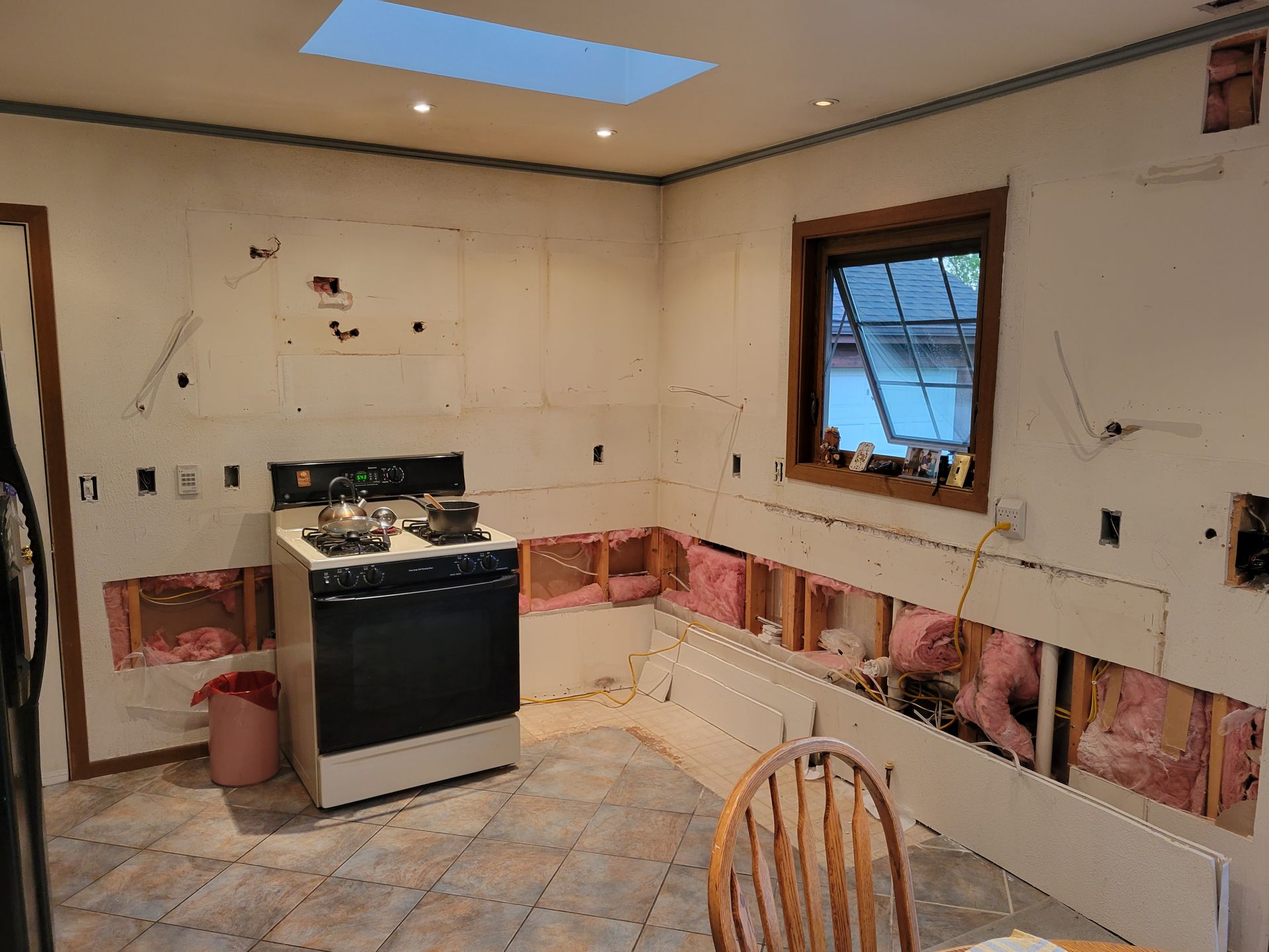 Damaged kitchen with torn walls, exposed insulation, and a stove beside a window under renovation.