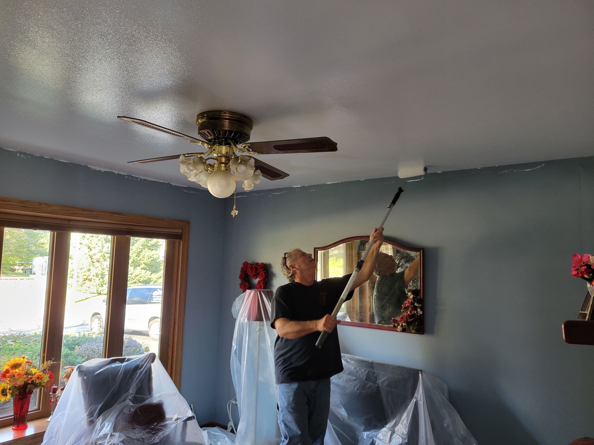 Person painting a blue wall in a living room with a ceiling fan and large window