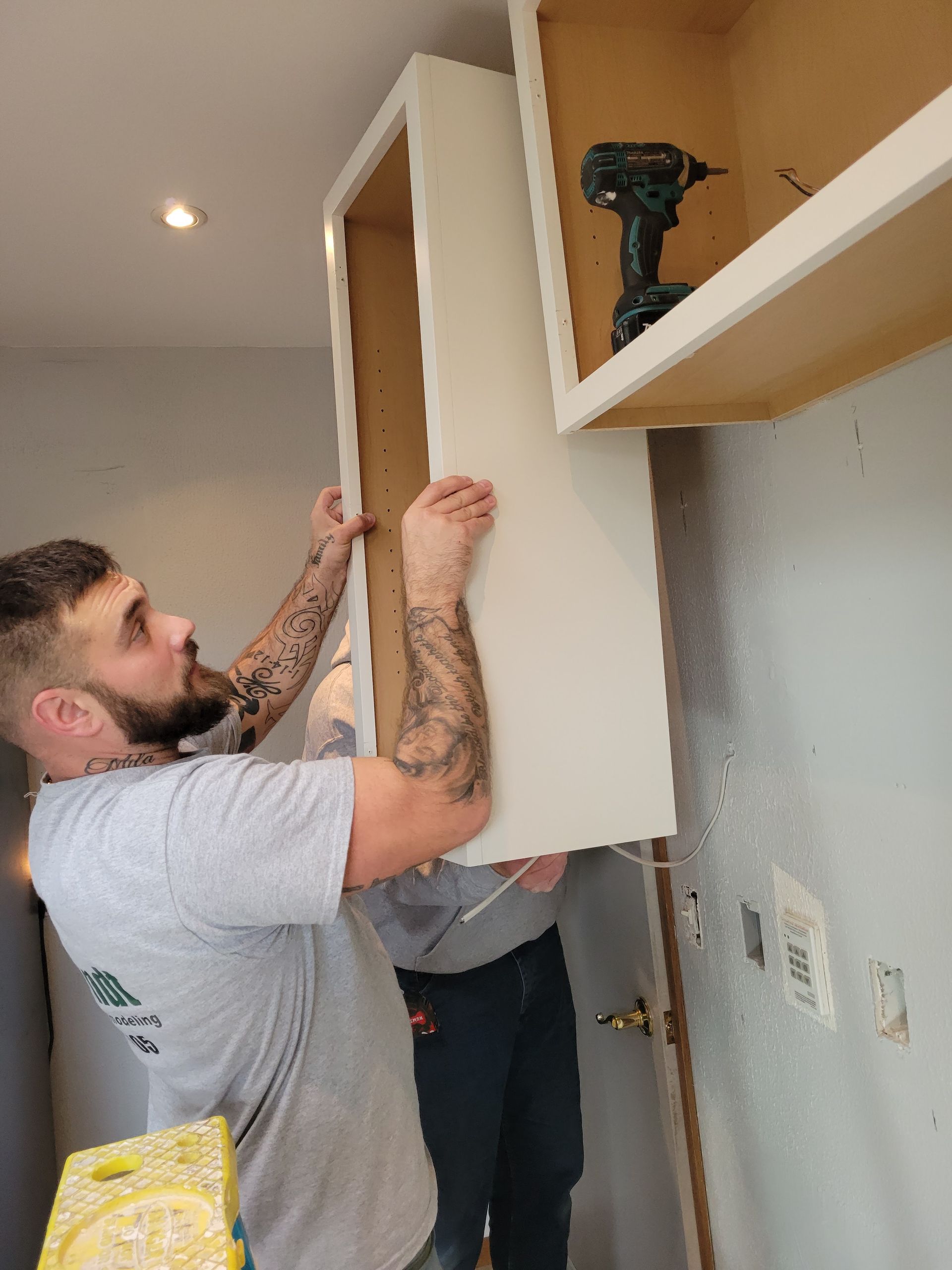 Two workers installing a white cabinet shelf in a room, one holding it in place near a wall outlet.