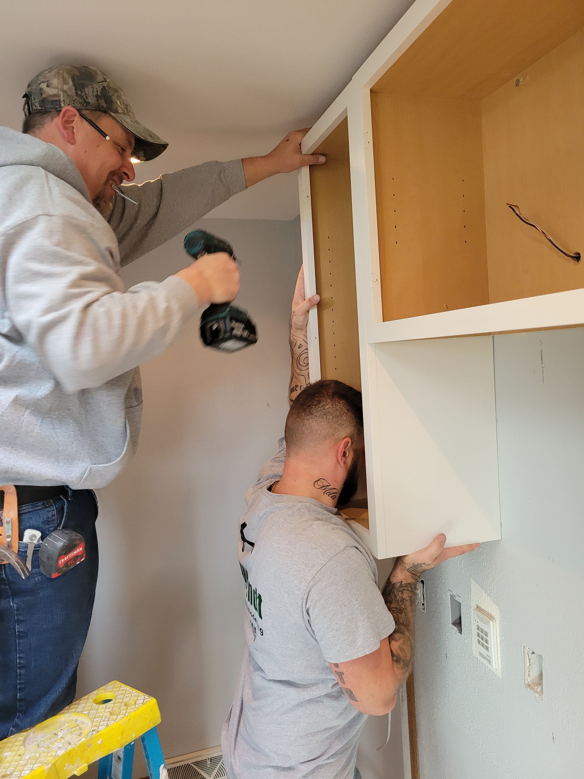Two workers installing white cabinets in a kitchen, one using a drill on a step stool.