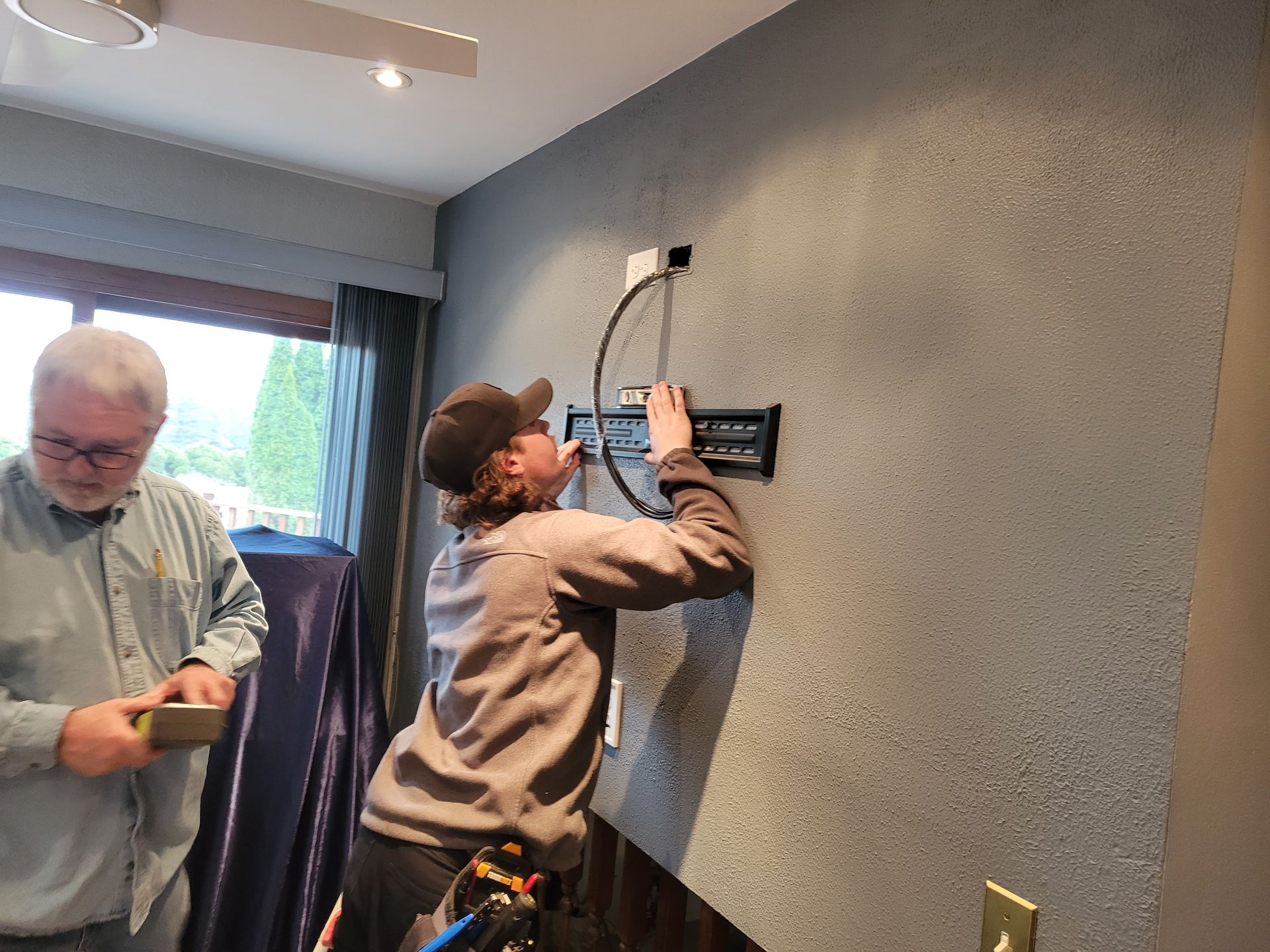 Two people mounting a wall device in a room, one holding it while the other works on the wiring.