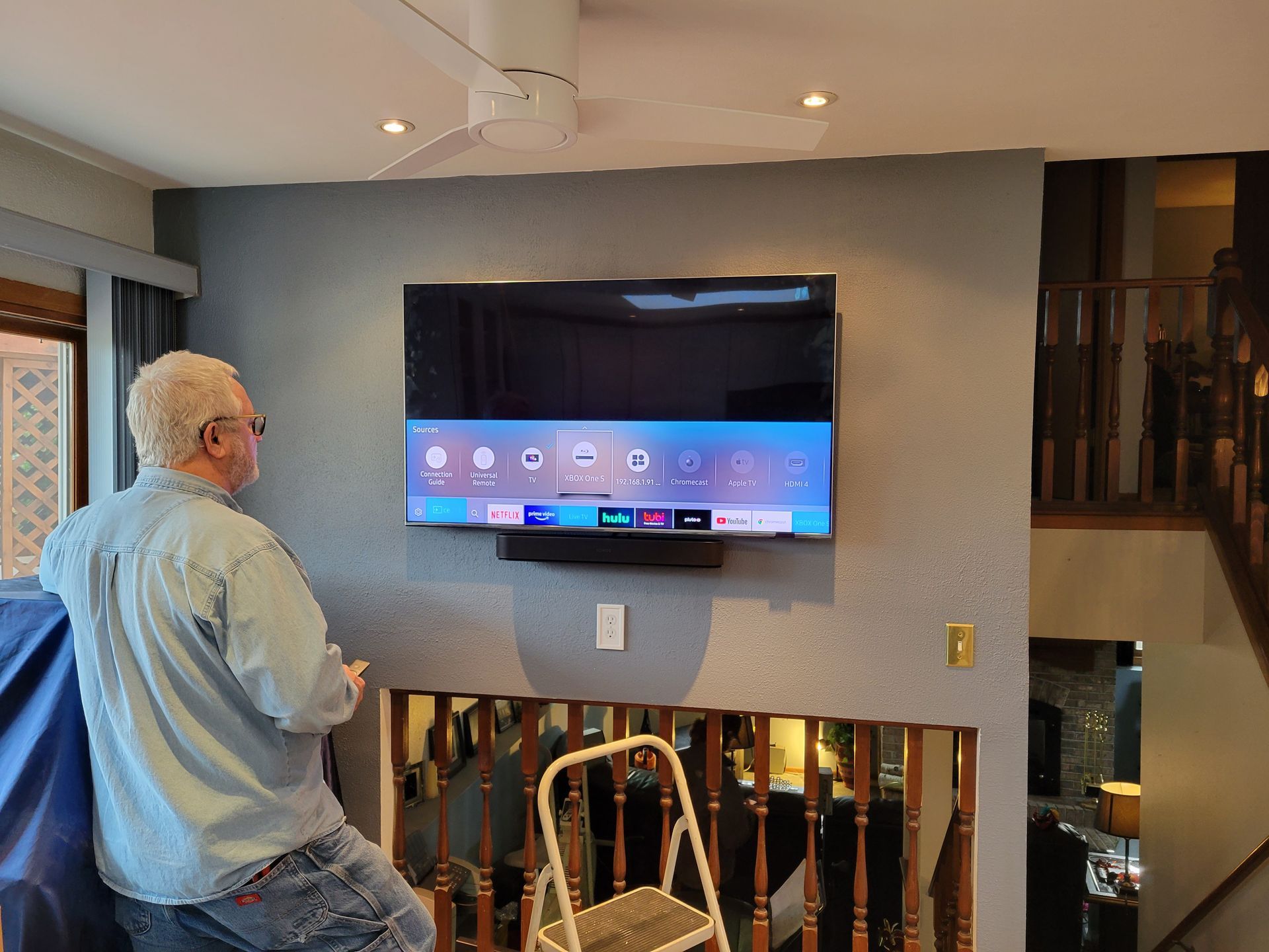 Man watching a wall-mounted TV in a home living room with ceiling lights and a staircase nearby