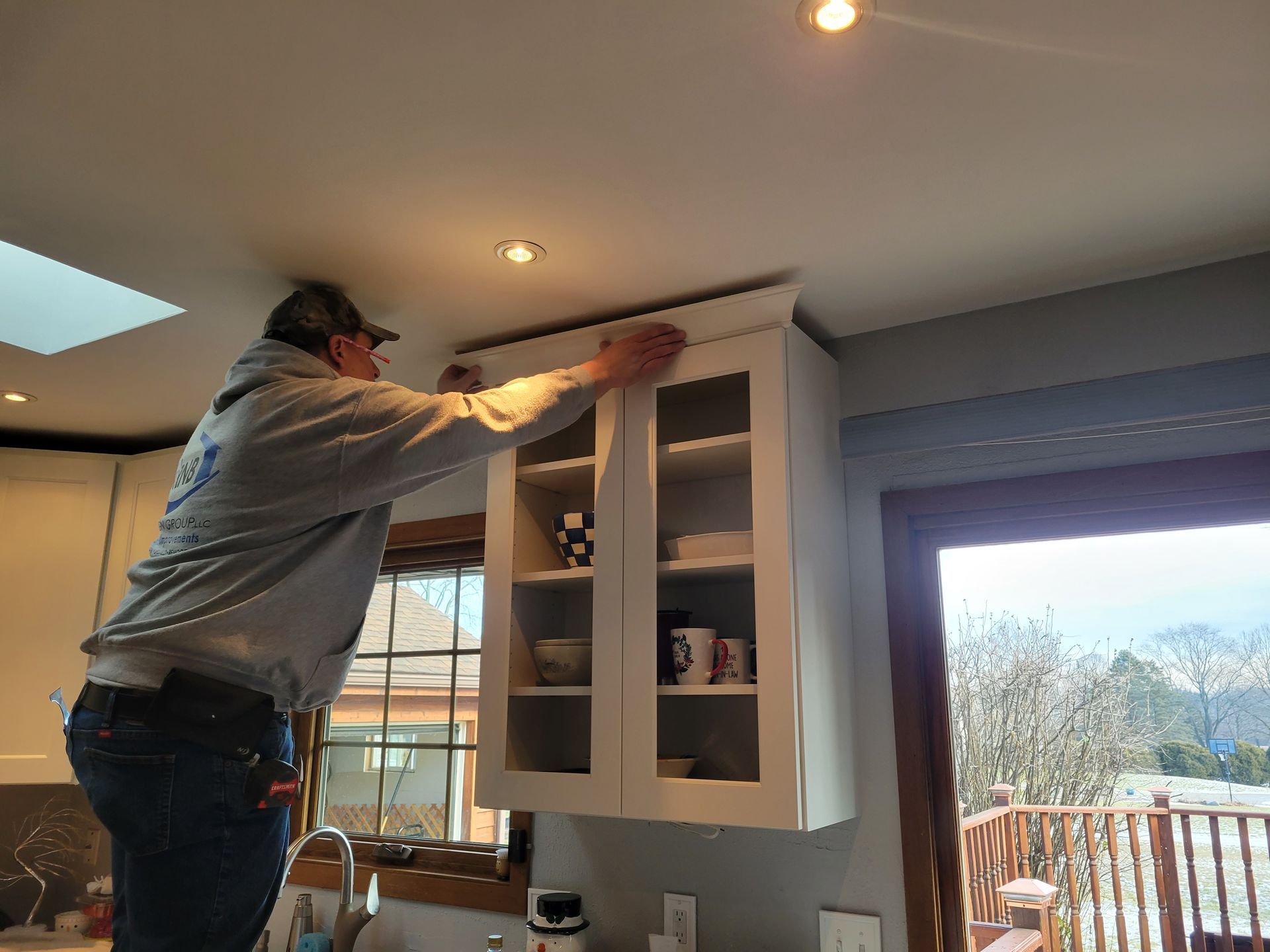 Person installing a white kitchen cabinet near a window, using a ladder in a bright room