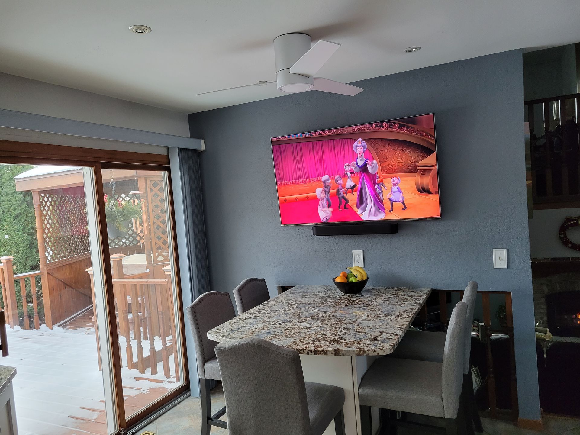 Dining area with granite table, gray chairs, and wall-mounted TV showing a colorful scene near sliding glass doors